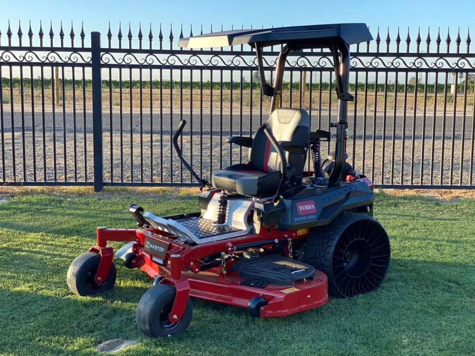 A red Toro Z Master zero-turn riding lawn mower on a grassy lawn in front of a black metal fence and an open field under a clear blue sky.