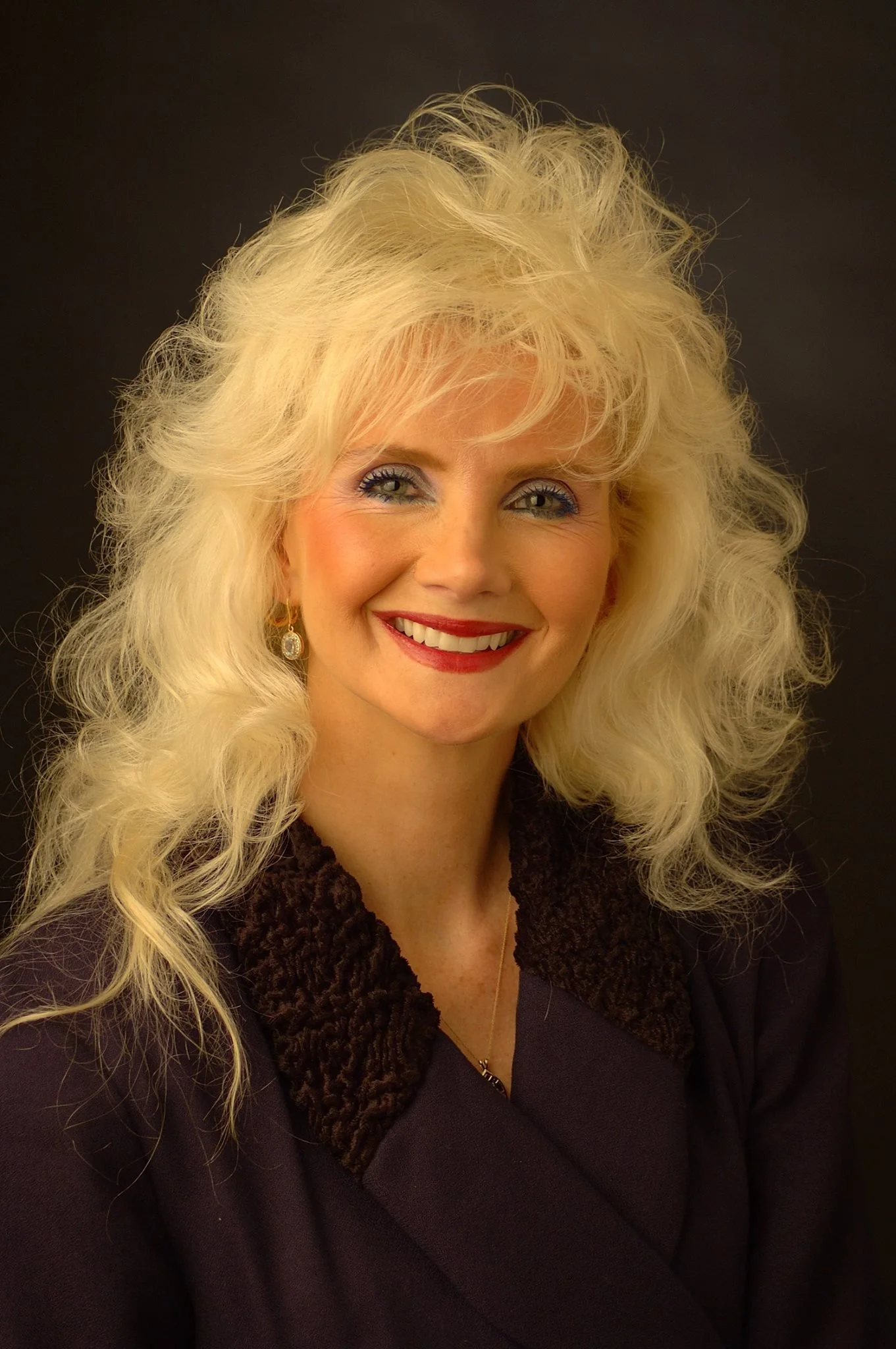 A woman with long blonde curly hair smiling, wearing earrings, a dark coat with a fur collar, and a gold necklace, against a black background.