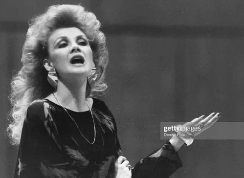 A woman with voluminous curly hair speaking or singing with her eyes closed and one hand raised.