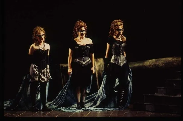 Three women in vintage-style dresses walking on a stage with a dark background.