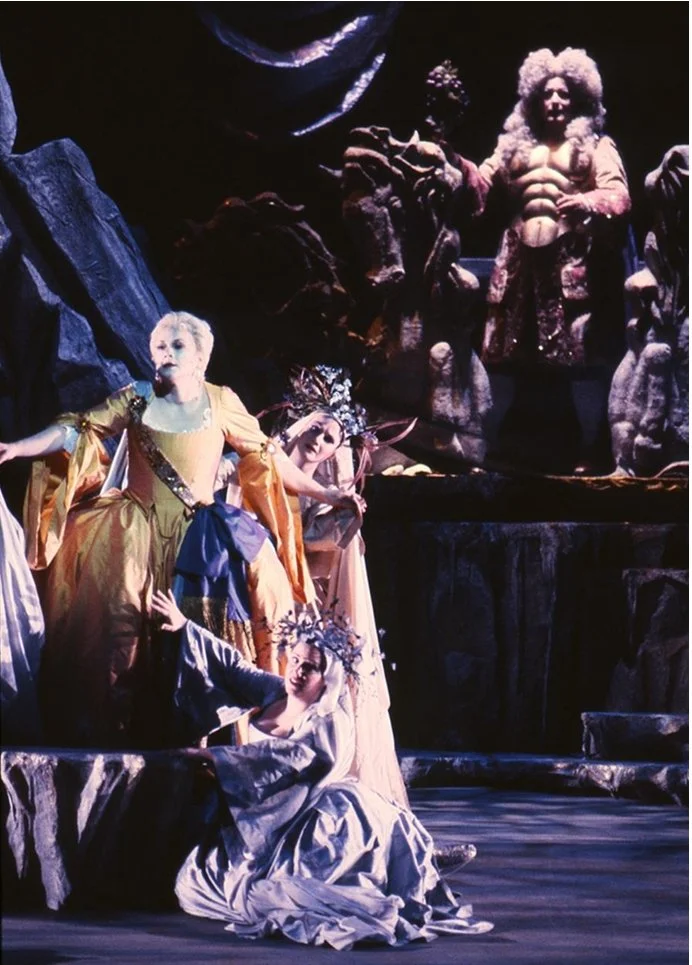 Actors dressed in elaborate costumes perform on stage, with a dark and rocky background, some wearing wigs and crowns, suggesting a theatrical or operatic scene.