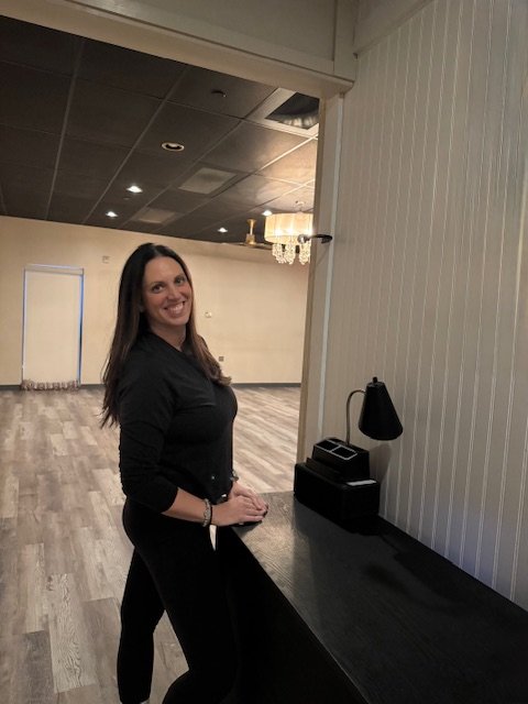 A woman in black clothing standing in a spacious room with wooden floor, white-paneled wall, and chandelier lighting, smiling at the camera.