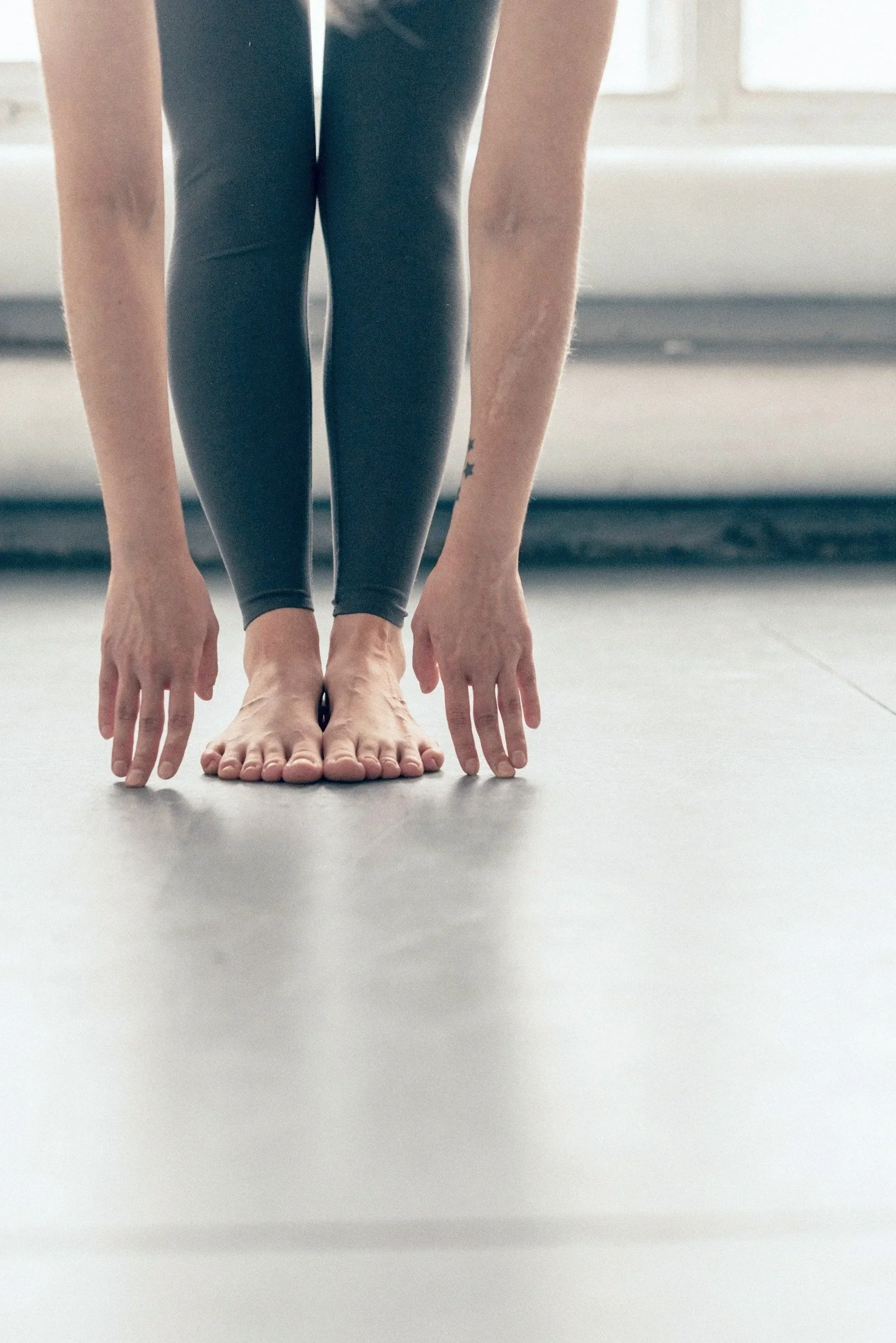 A person in black leggings and a tattoo on their arm is standing on a gray floor in a yoga pose with their hands and feet on the ground.