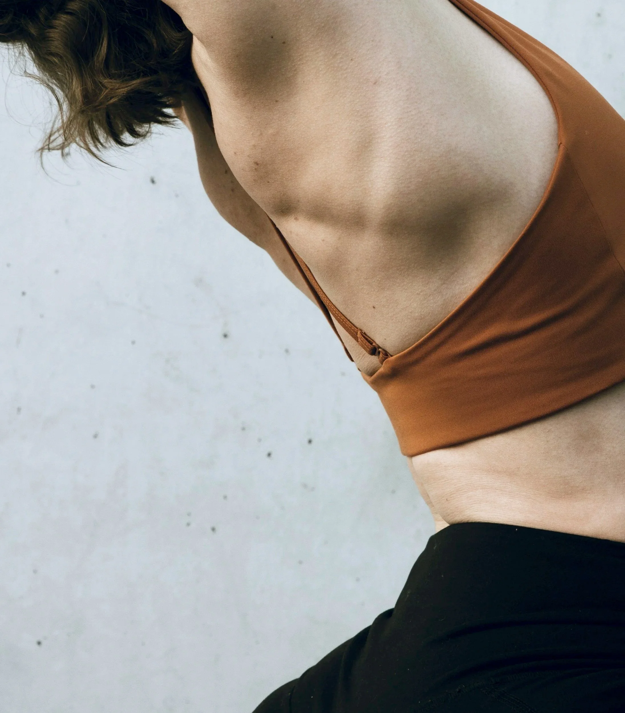 A person wearing a brown tank top and black leggings, leaning forward against a plain gray wall.