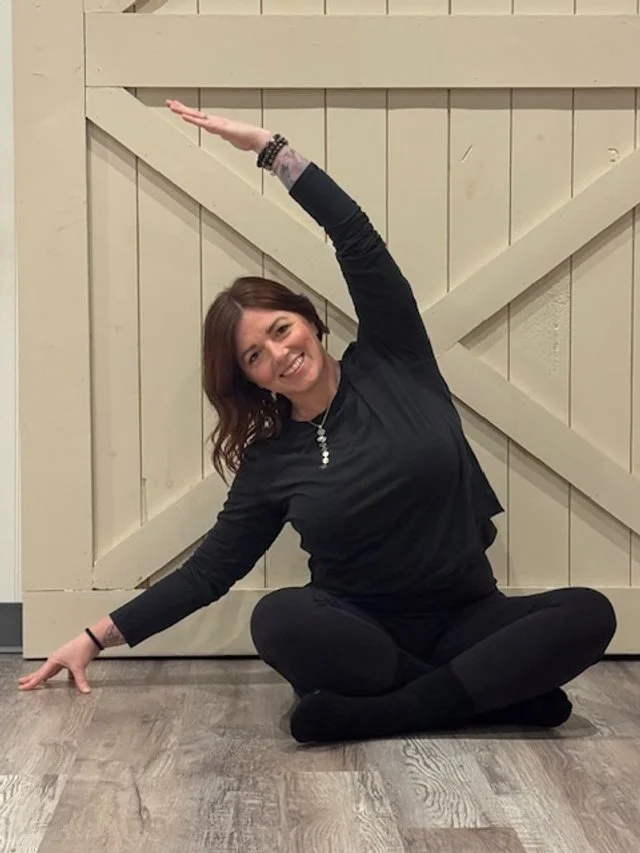 A woman with brown hair sits cross-legged on the floor with a wood-paneled wall behind her. She is smiling and stretching, with her left arm reaching upwards and her right arm reaching sideways.