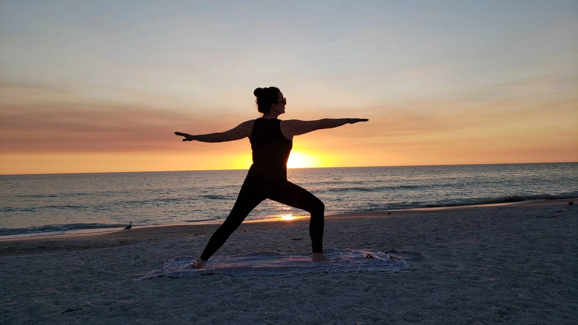 A woman practicing yoga on the beach during sunset, standing in warrior pose with arms extended and facing the ocean.