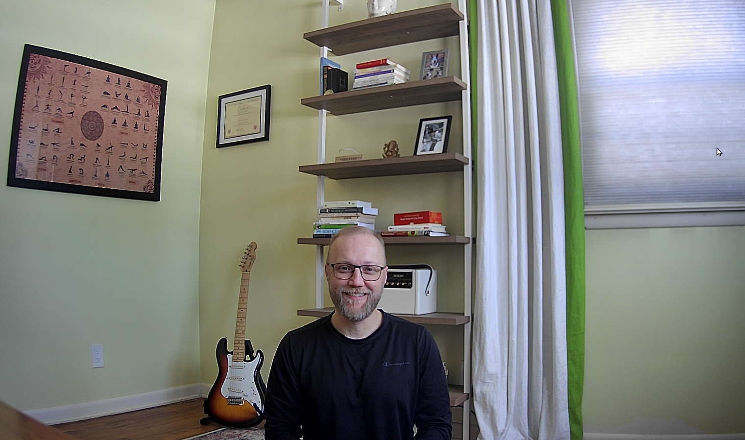 A man with glasses, a beard, and a bald head smiling in a room with a green wall. There is a bookshelf behind him with books and decorative items, and a framed certificate on the wall. A sunburst electric guitar is leaning against the wall near the floor, and there is a window with white curtains and green accents on the right side.