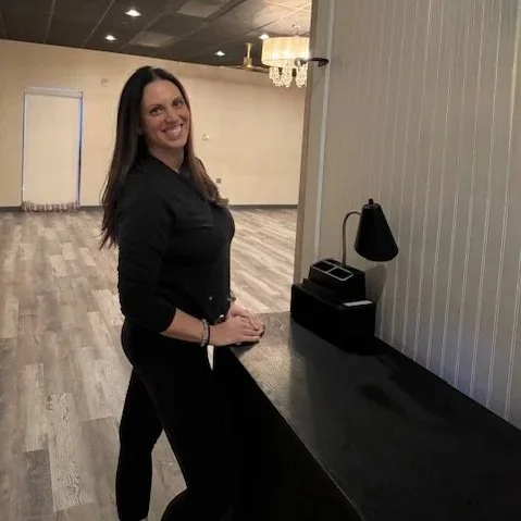 A woman with long dark hair smiling and standing at a black counter in a spacious room with wooden flooring and a chandelier.