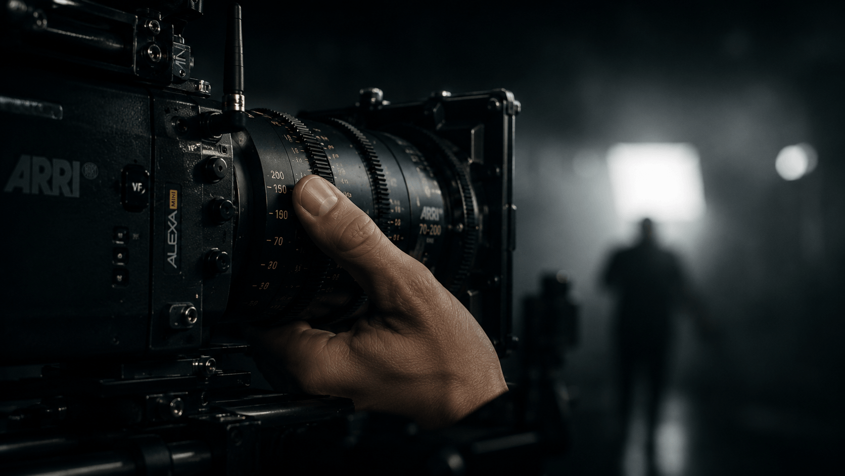 Persona ajustando el enfoque de una cámara de cine en un set oscuro, con una figura borrosa en el fondo.