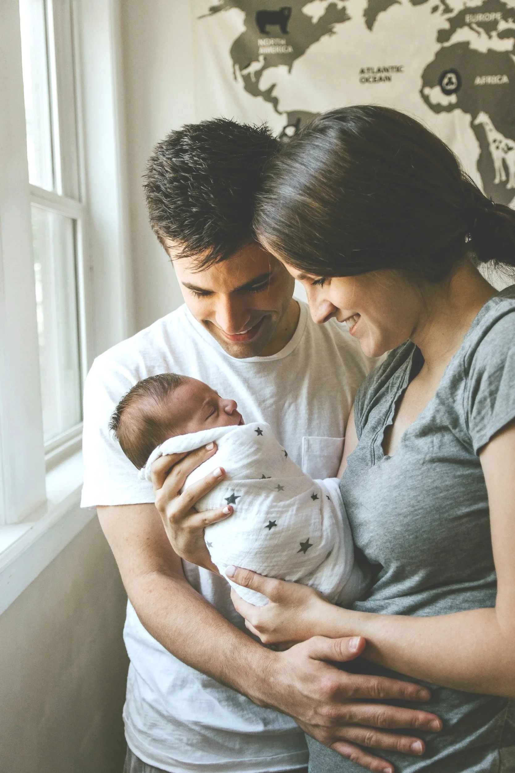 A smiling couple holding a newborn baby wrapped in a star-printed blanket, standing near a window with a world map on the wall behind them.