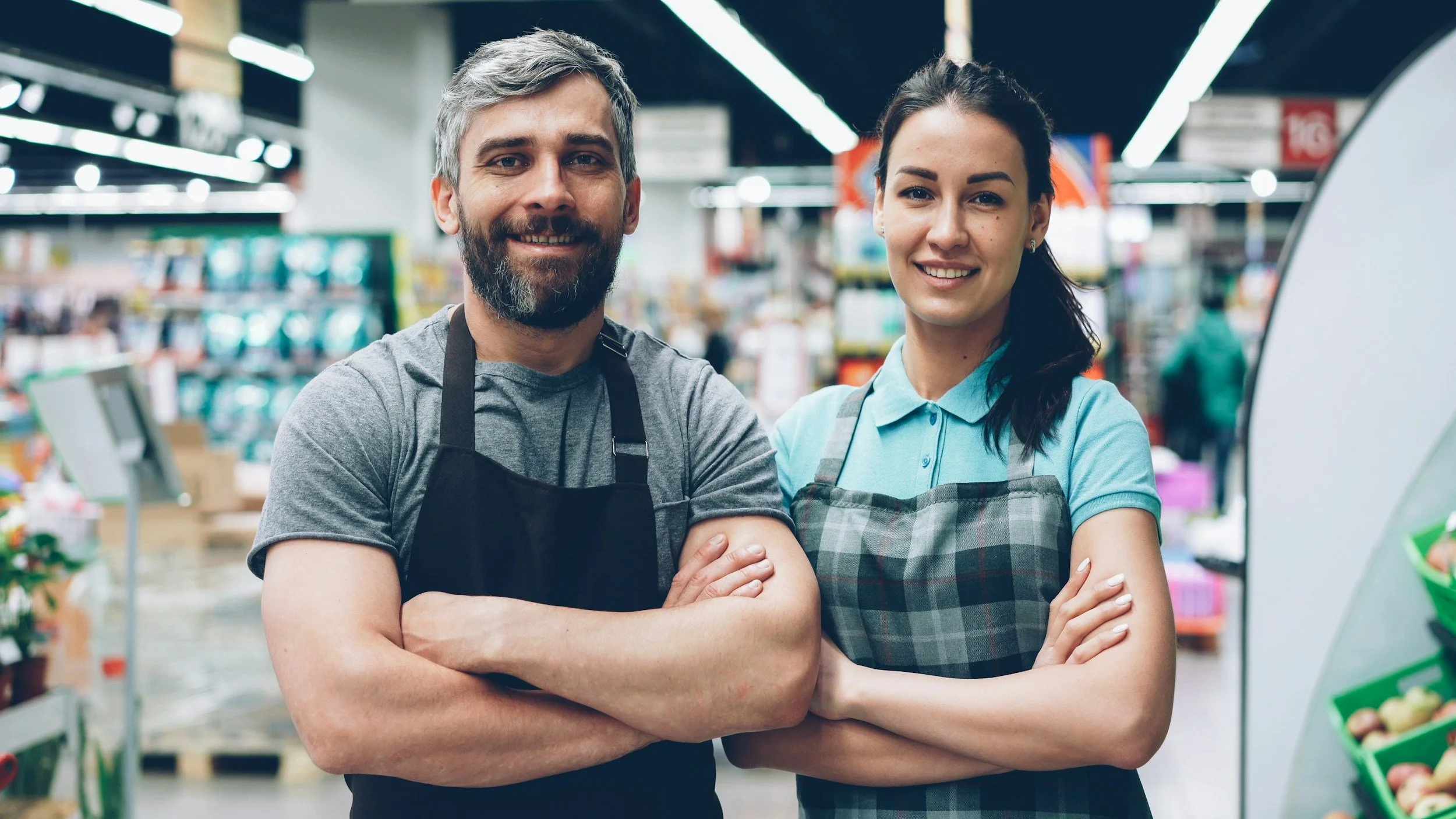 Two smiling employees, a man and a woman, standing side by side with arms crossed inside a grocery store, wearing aprons.