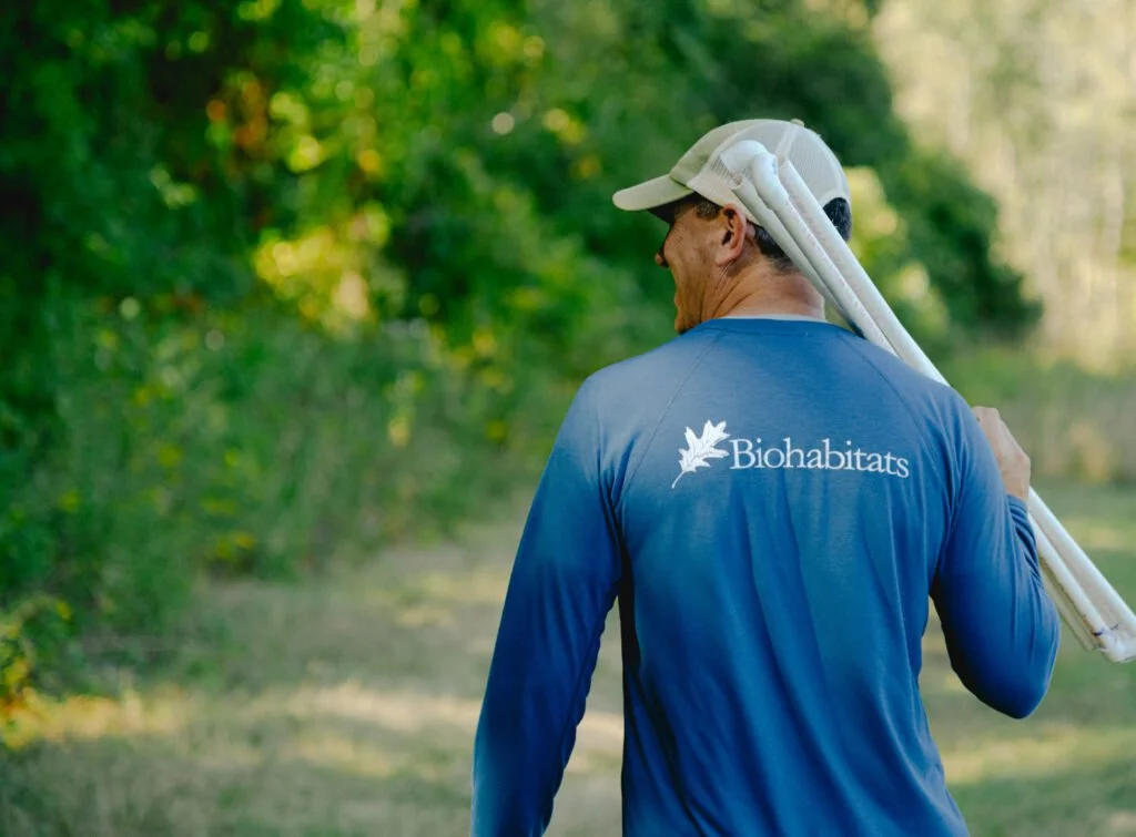 Man with a blue Biohabitats shirt carrying white pipes over his shoulder in a green outdoor setting.