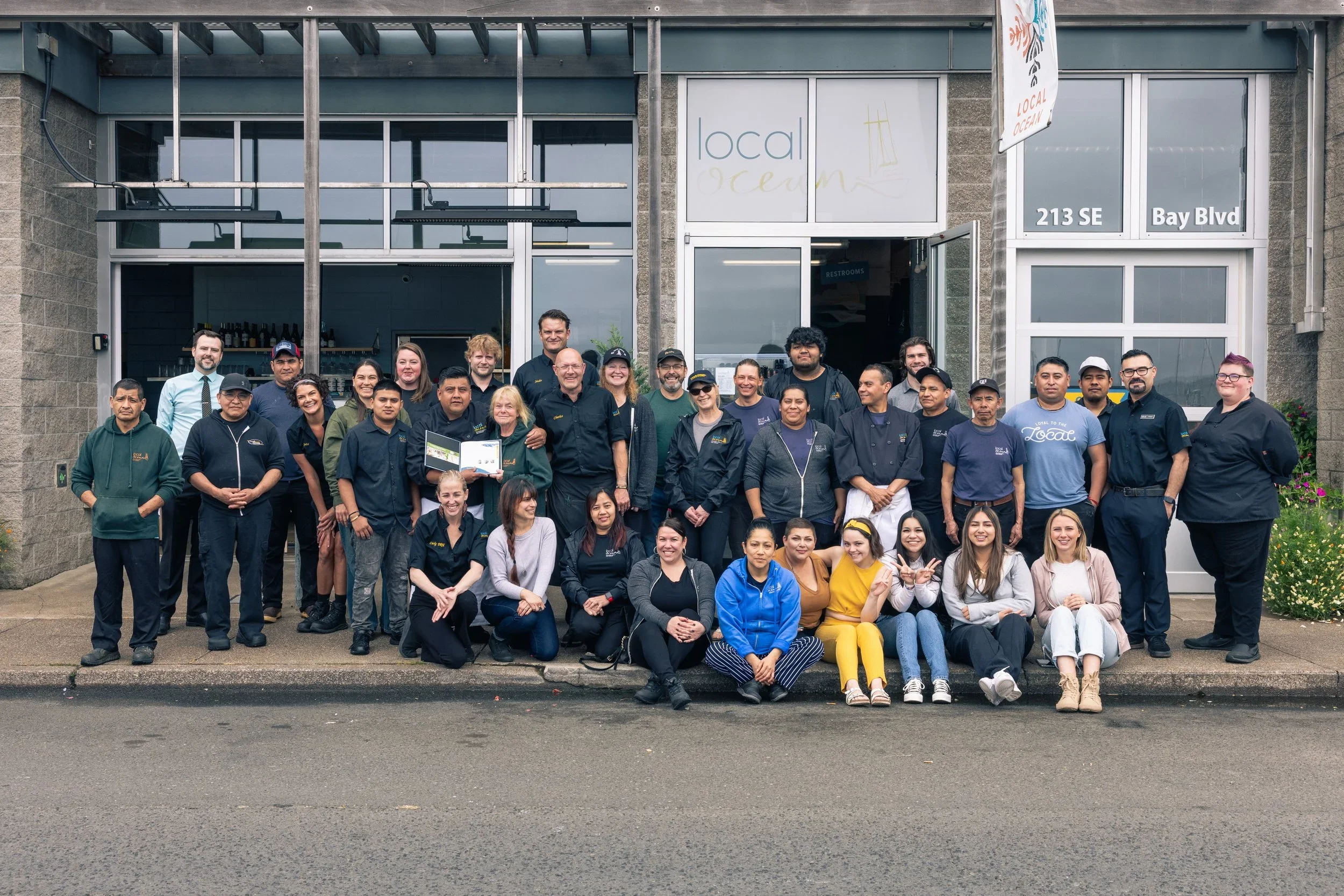 Group of people posing in front of a building with a sign reading 'local ocean' and an address '213 SE Bay Blvd'
