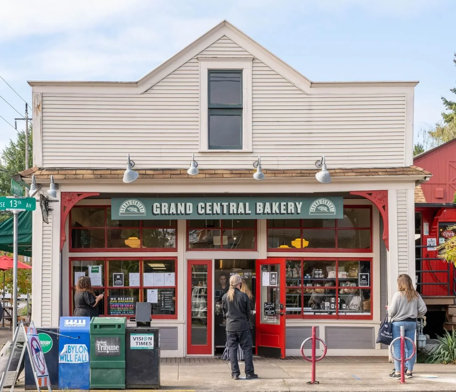 Exterior view of Grand Central Bakery storefront with three people standing outside and a street sign noting 13th Avenue. The bakery has a green sign, red window frames, and a red door.
