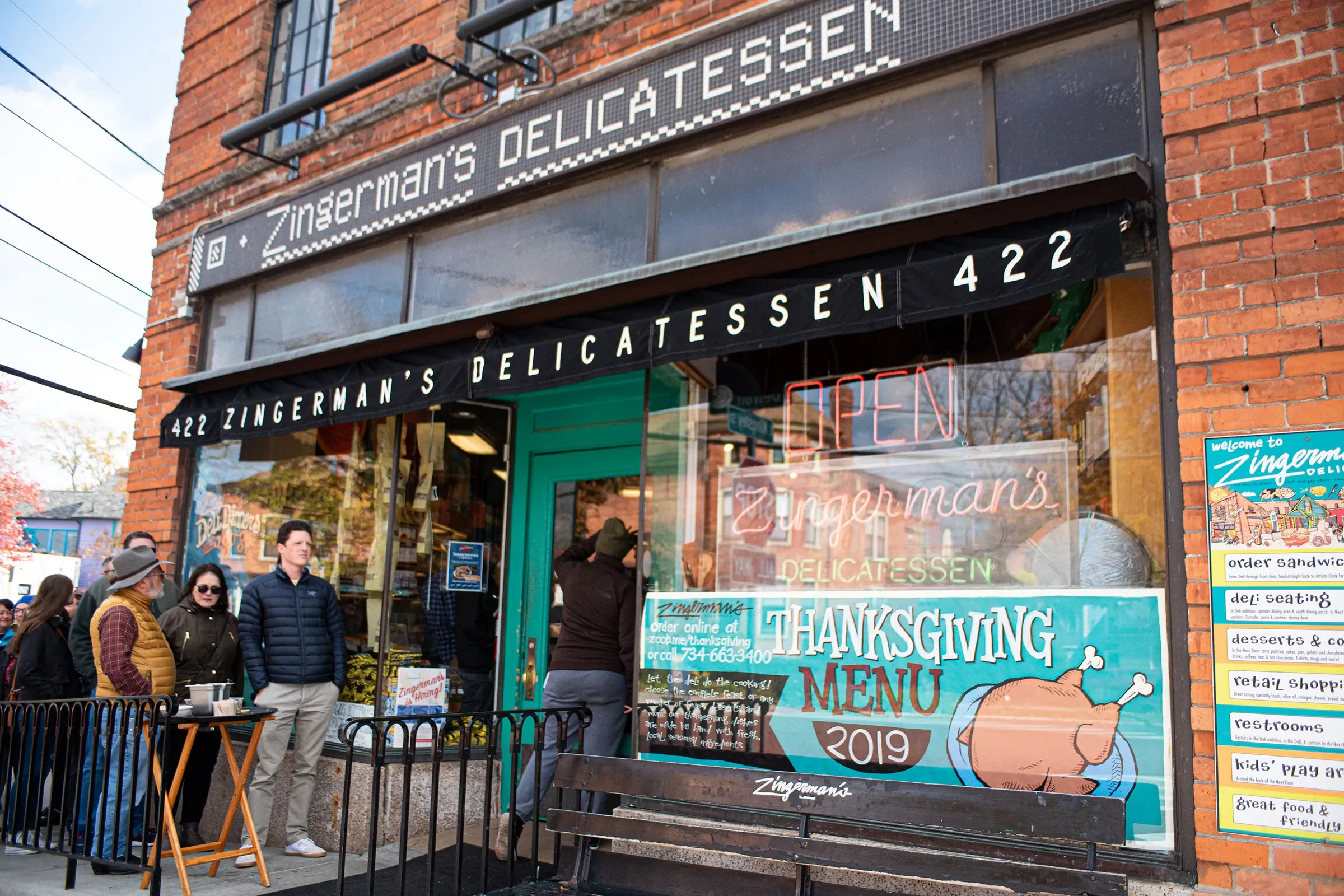 People standing outside Zingerman's Delicatessen, a brick storefront with a black awning, neon 'OPEN' sign, and colorful Thanksgiving menu poster.