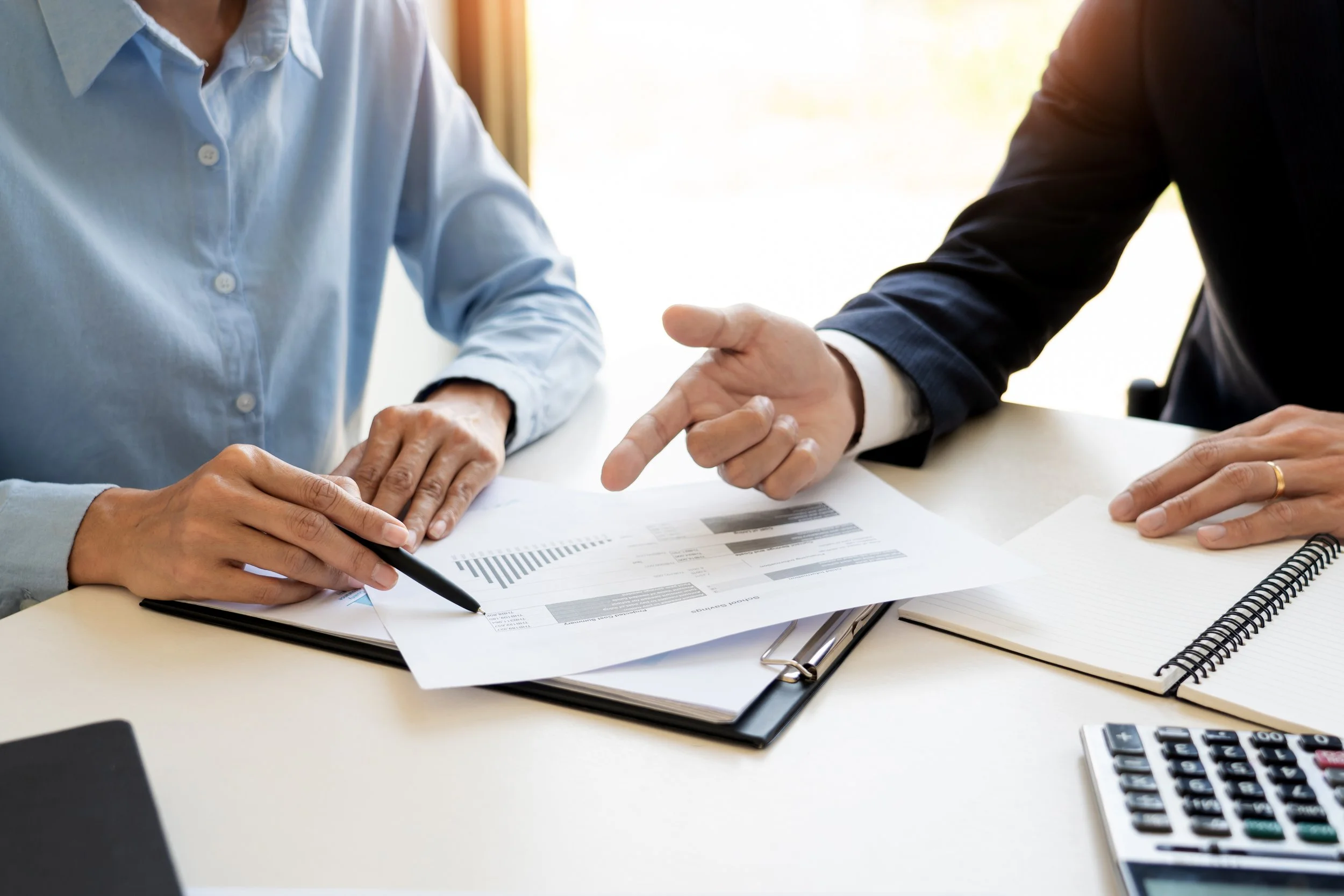 Two people in business attire discussing documents on a desk, with a pen, notebook, calculator, and financial papers visible.