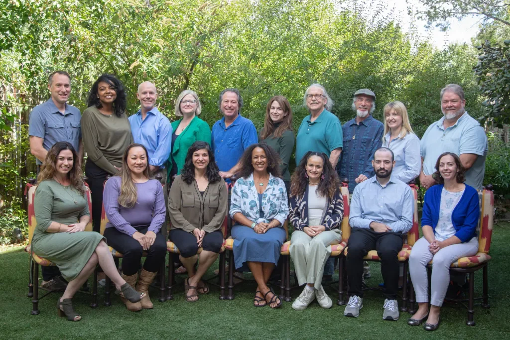 Group of 16 people posing outdoors in a garden, some sitting on chairs and others standing behind, surrounded by green trees and bushes.