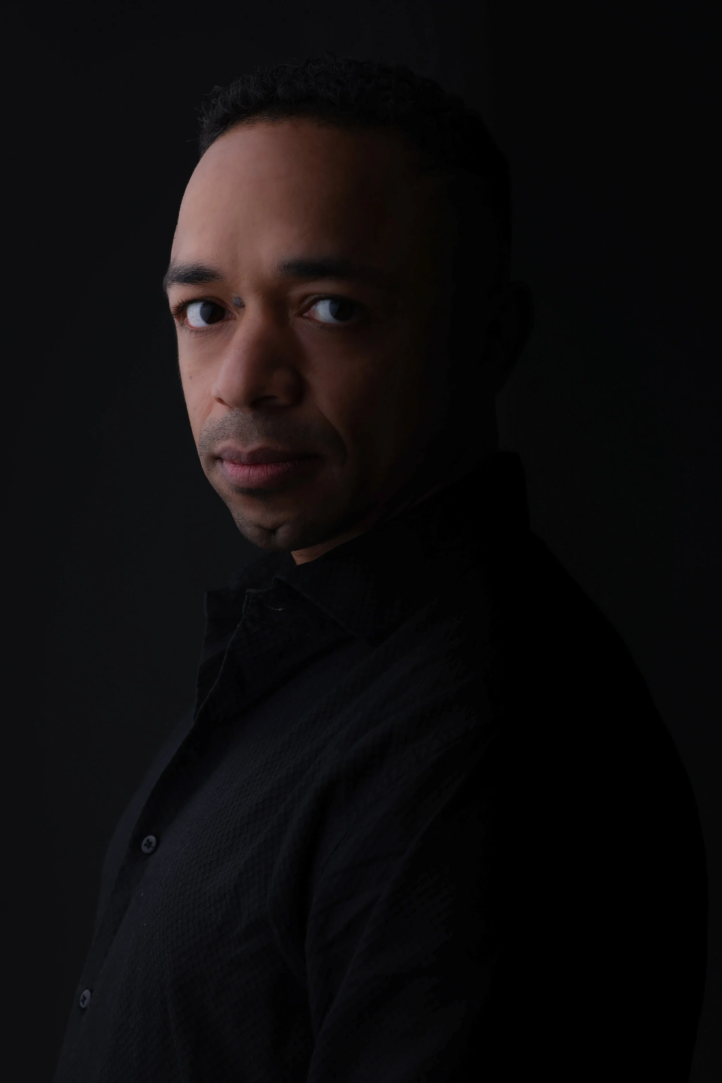 Portrait of dj xearo with dark curly hair, wearing a black shirt, against a dark background.