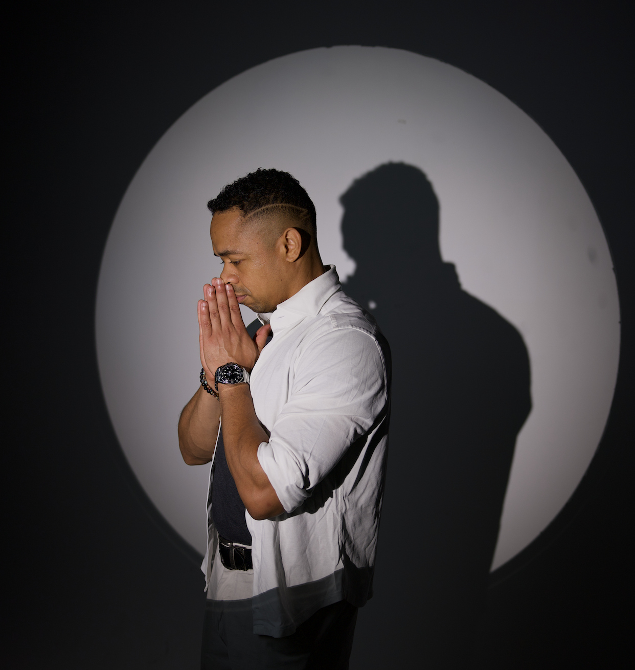 DJ Xearo with short curly hair and a fade haircut praying with his hands together, standing in front of a spotlight that casts a shadow of himself on the wall.