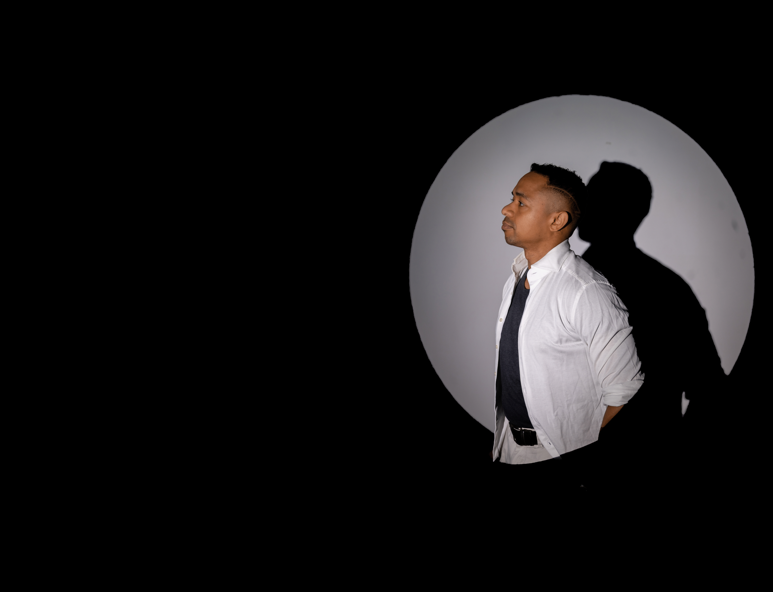 DJ xearo in a white shirt and dark t-shirt stands in profile within a circular spotlight,; his shadow cast onto a light gray background.
