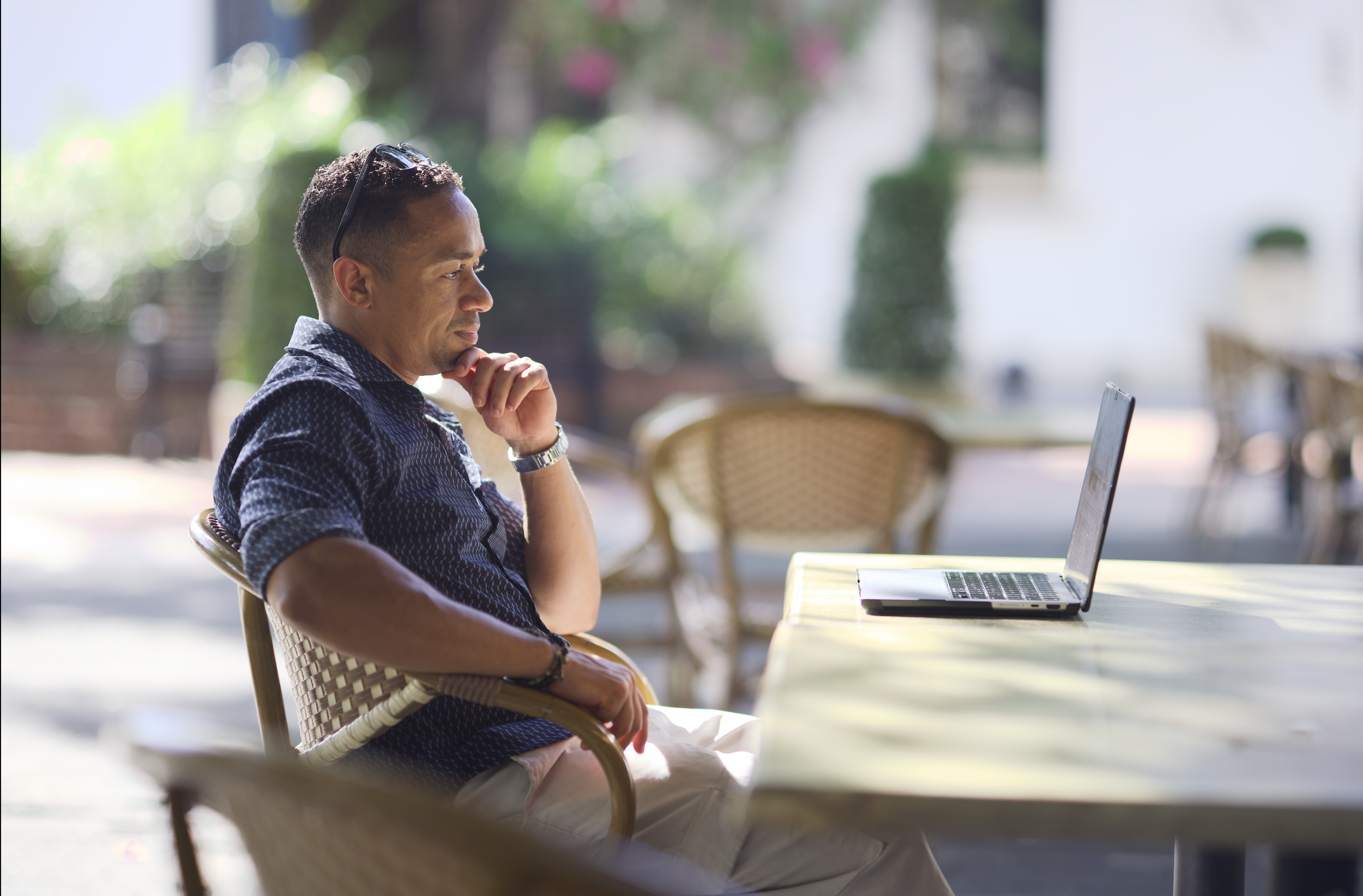 Carlos Manuel sitting outdoors at a table, looking at a laptop with a thoughtful expression.