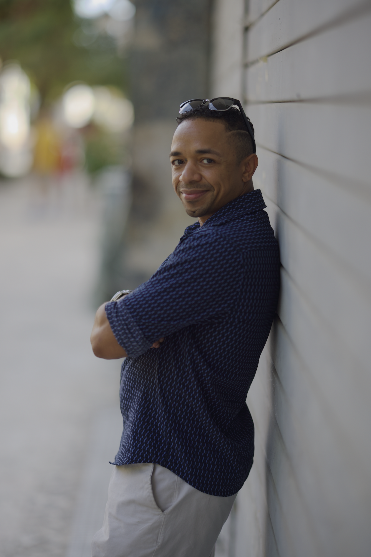 Carlos Manuel wearing a dark blue patterned shirt and light-colored pants leaning against a light-colored wall outdoors, smiling at the camera with sunglasses resting on his head.