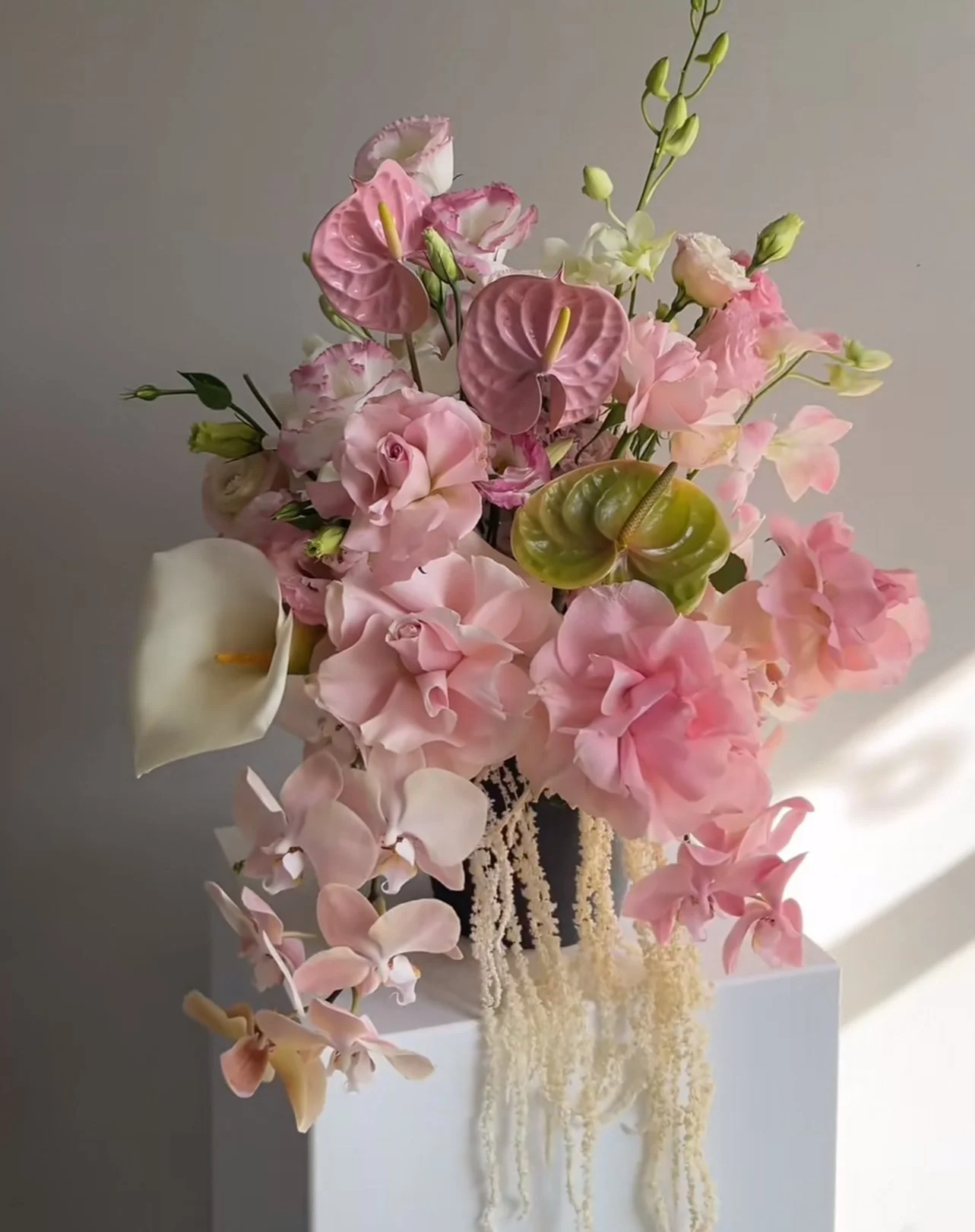 A floral arrangement with pink, white, and green flowers, including calla lilies, anthuriums, and other mixed blooms, displayed on a white pedestal against a neutral wall.