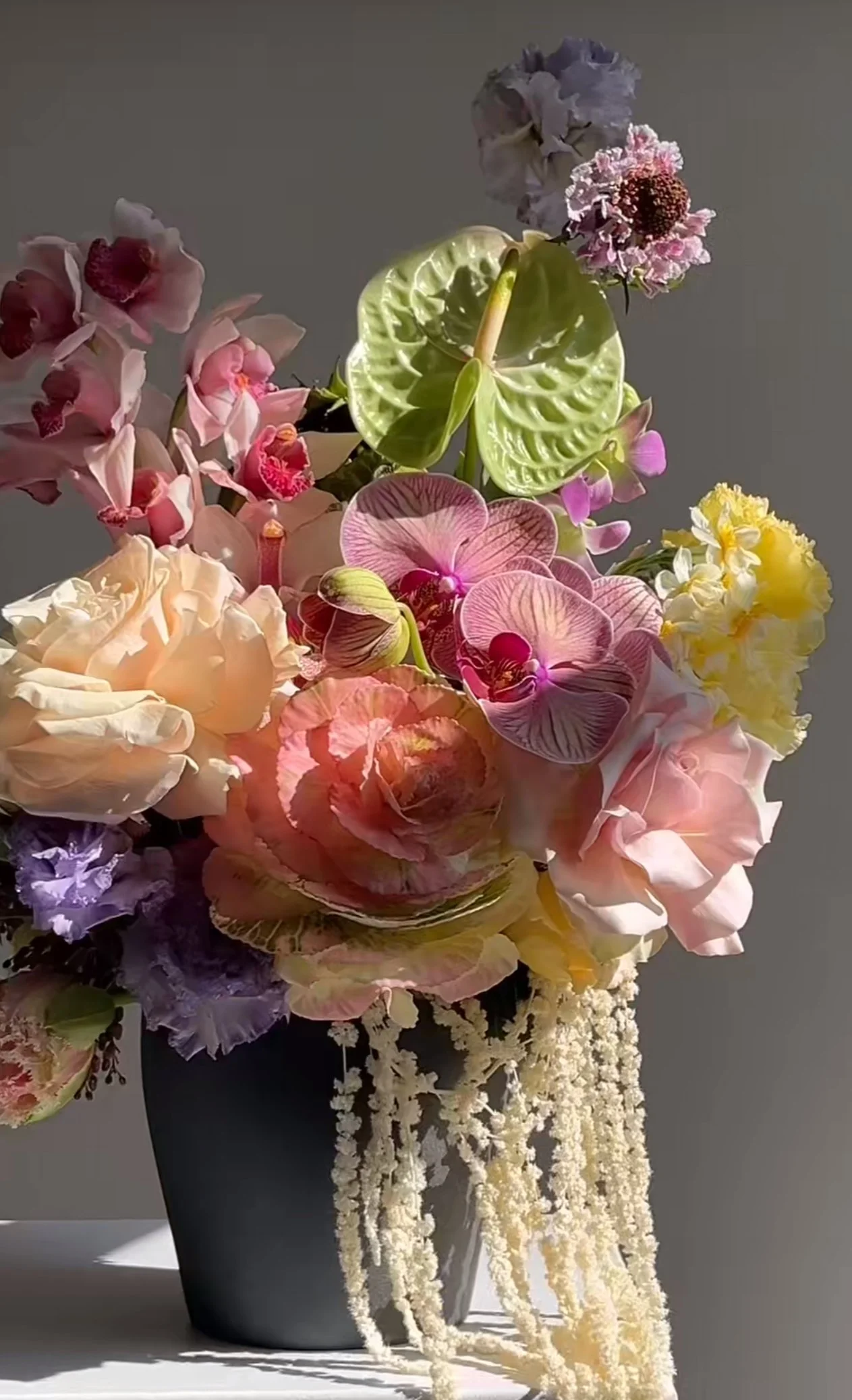 A mixed floral bouquet in a black vase, featuring various pink, purple, yellow, and white flowers including orchids and calla lilies, with cascading white flowers at the base.