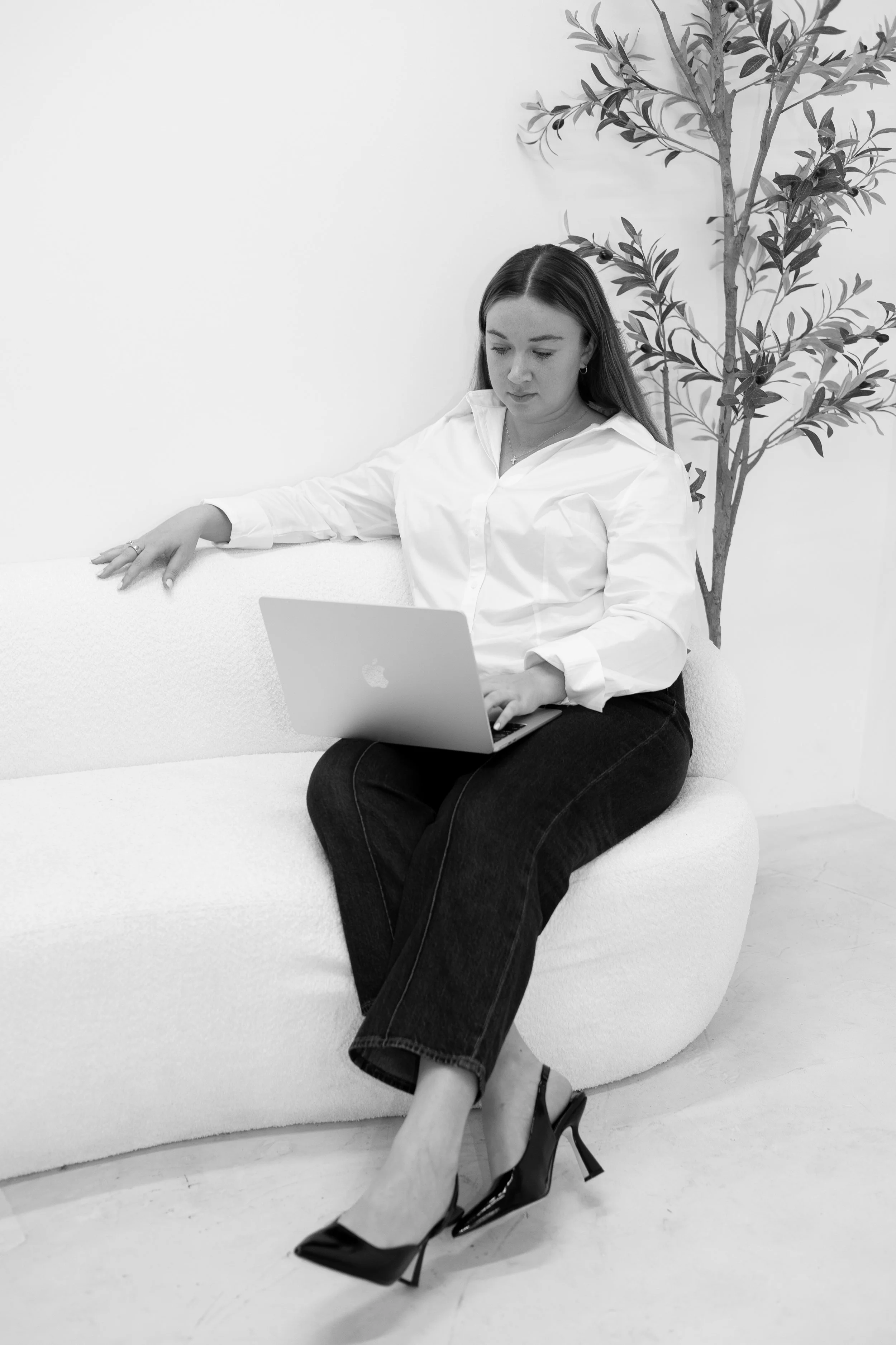A woman sitting on a white couch, working on a laptop, with a potted plant in the background.