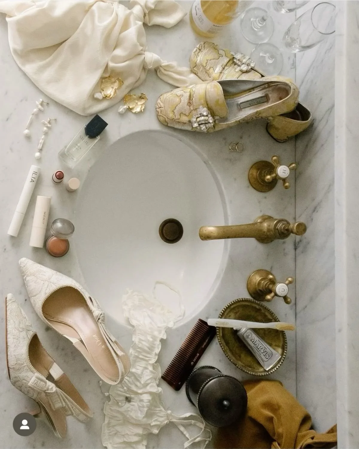 Bathroom sink with multiple personal items including patterned shoes, white clothes, makeup products, a comb, perfume, jewelry, glasses, and a pair of shoes on a marble countertop.