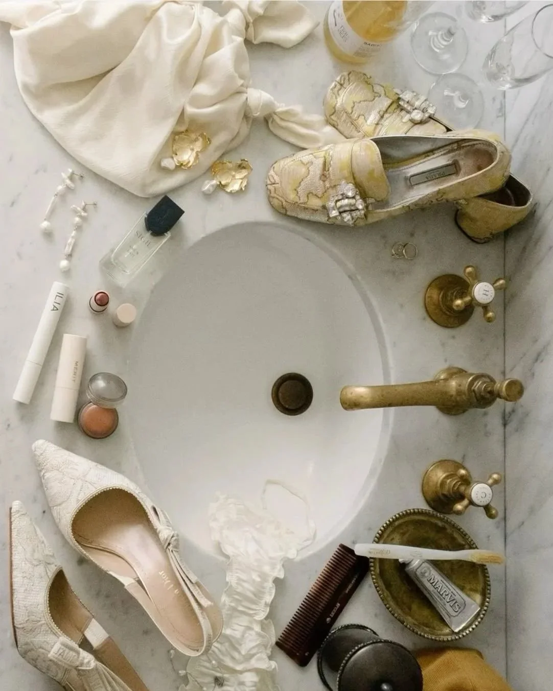 Objects on a bathroom sink surrounding an empty basin, including a pair of cream-colored embellished shoes, white pants, makeup items, jewelry, and perfume.