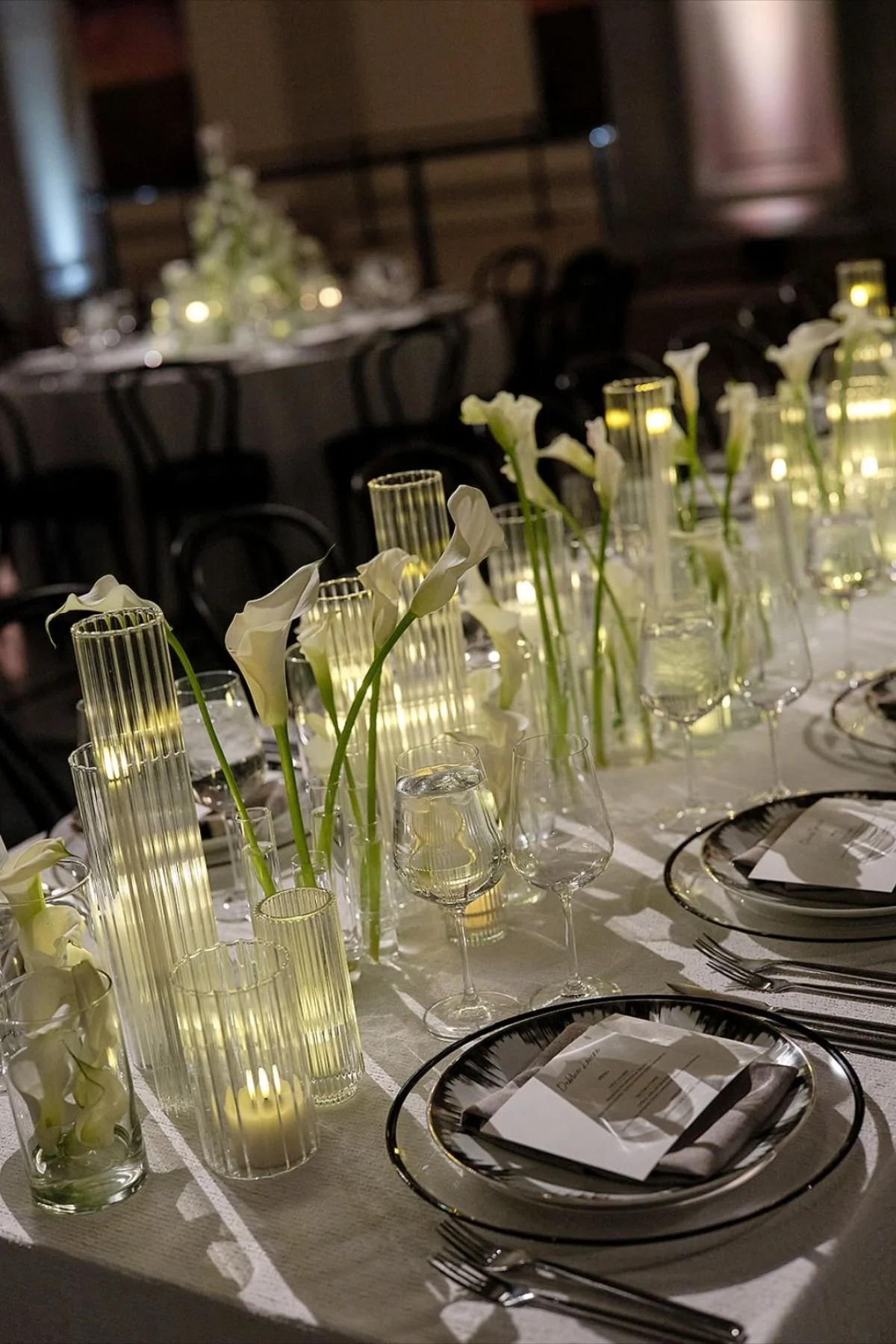 Elegant table setting with white calla lilies in tall vases, lit candles in glass holders, wine glasses, and black and gold-rimmed plates at a formal event.