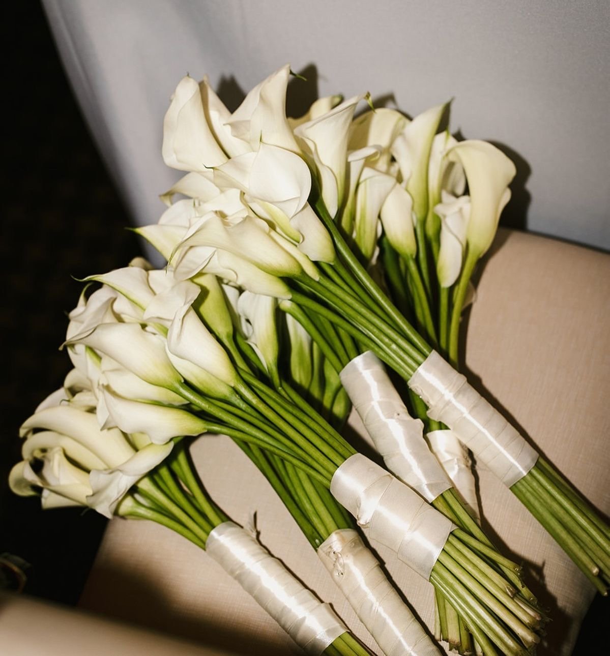 Two bunches of white calla lilies wrapped with white satin ribbon on a beige chair.