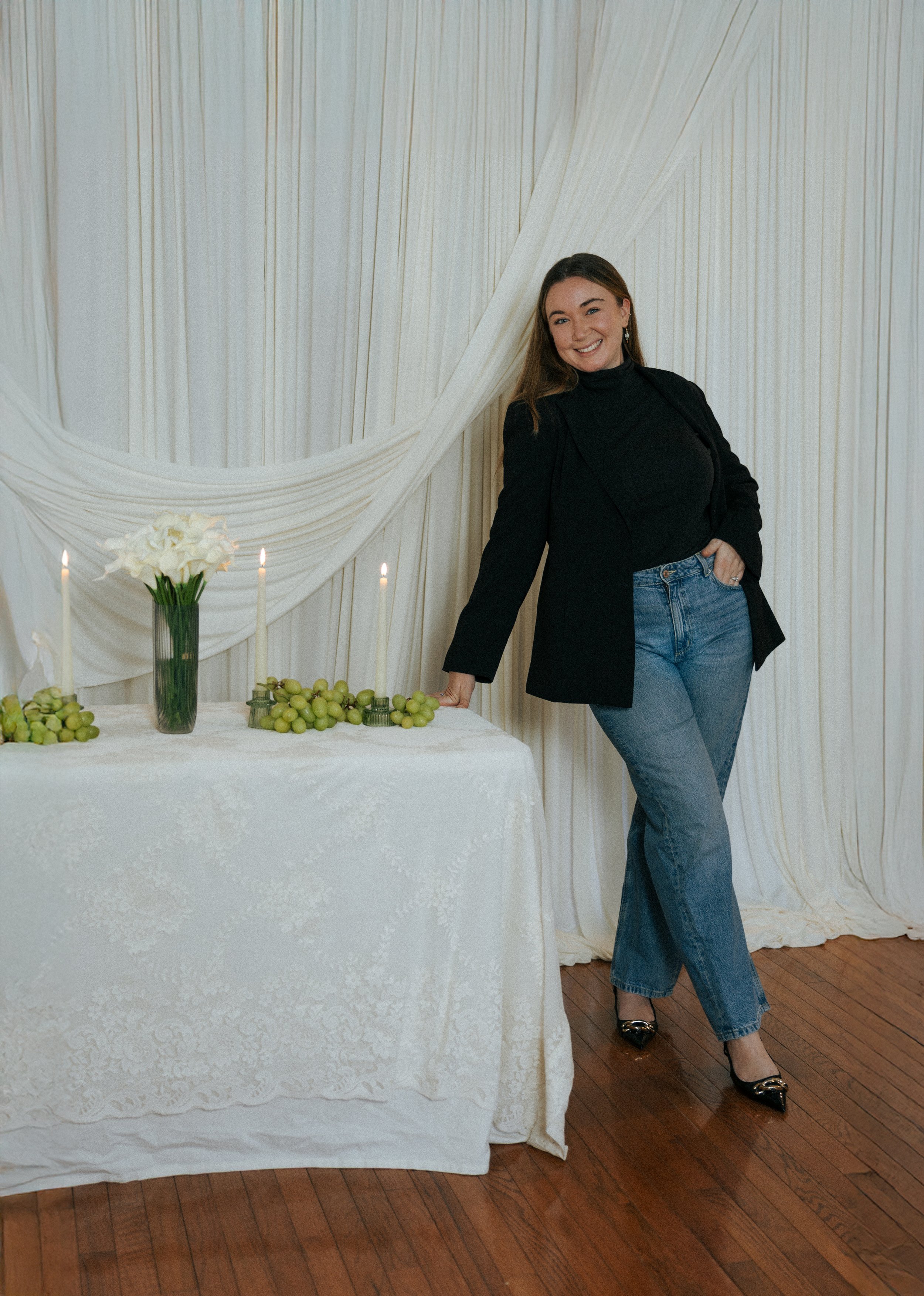A woman in a black blazer, black turtleneck, and blue jeans standing next to a table with white tablecloth, green grapes, white flowers, and lit candles against cream-colored draped curtains.
