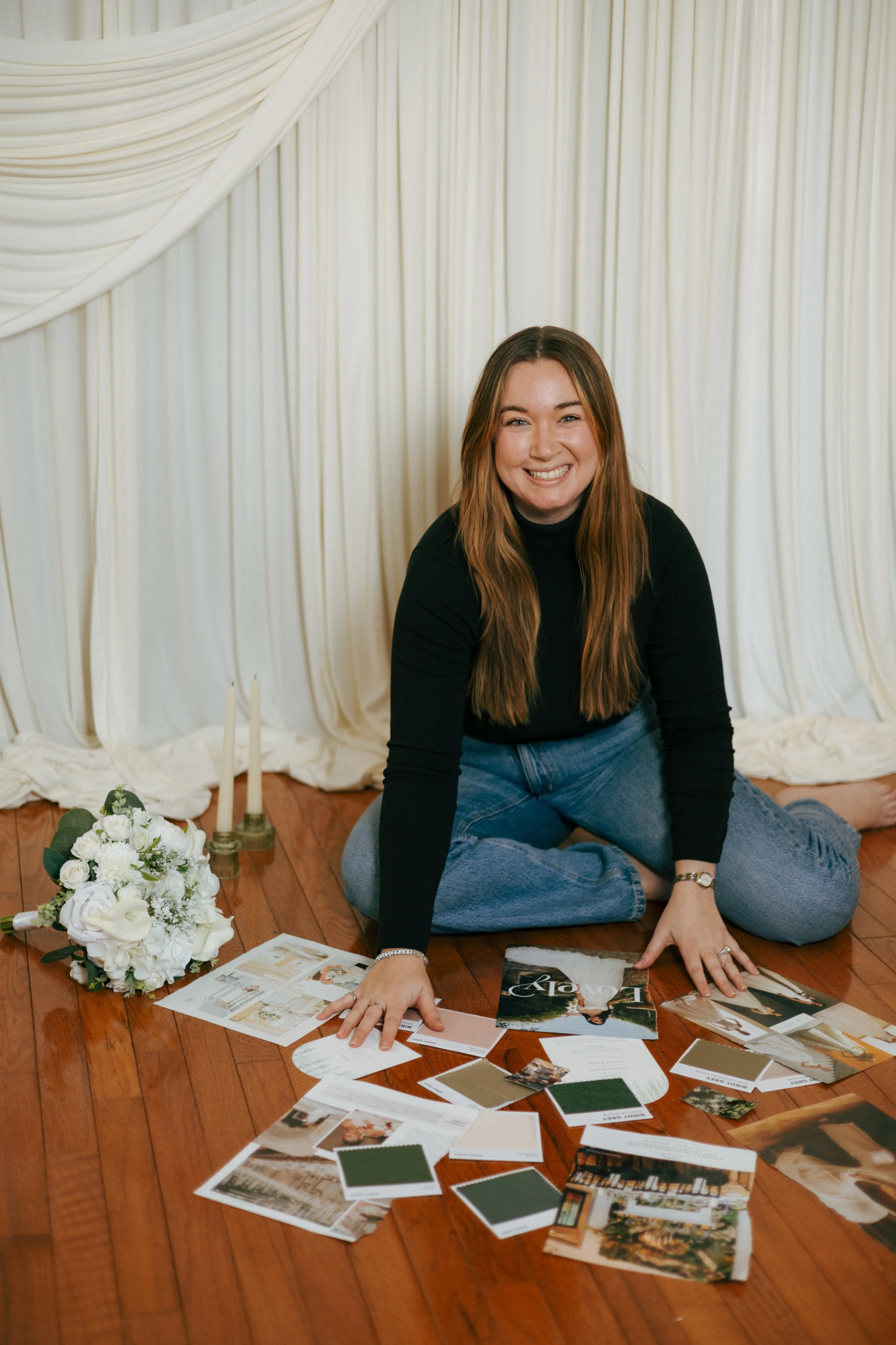 Woman sitting on wooden floor surrounded by wedding planning materials including photos, swatches, and wedding notes, with a bouquet of white flowers and two candlesticks nearby, in front of a cream-colored draped backdrop.