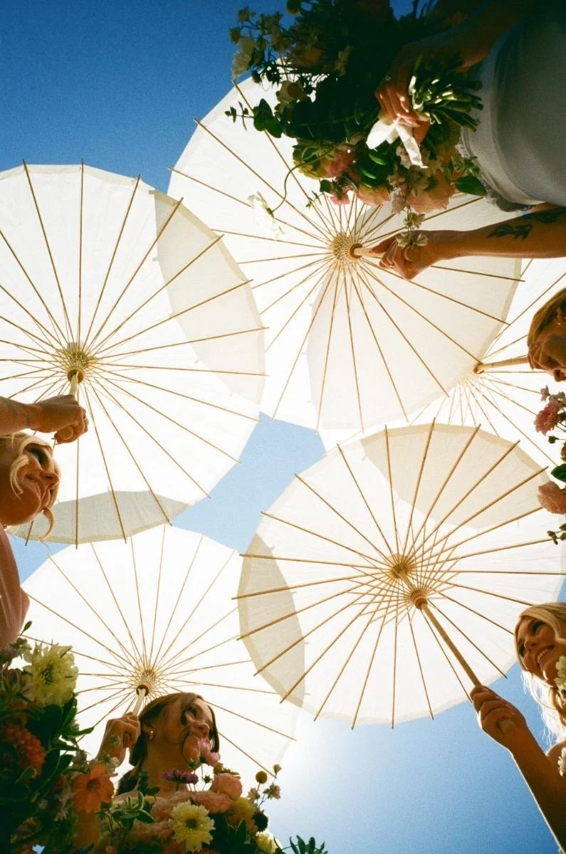 A group of women holding white parasols and flowers, seen from below against a blue sky.