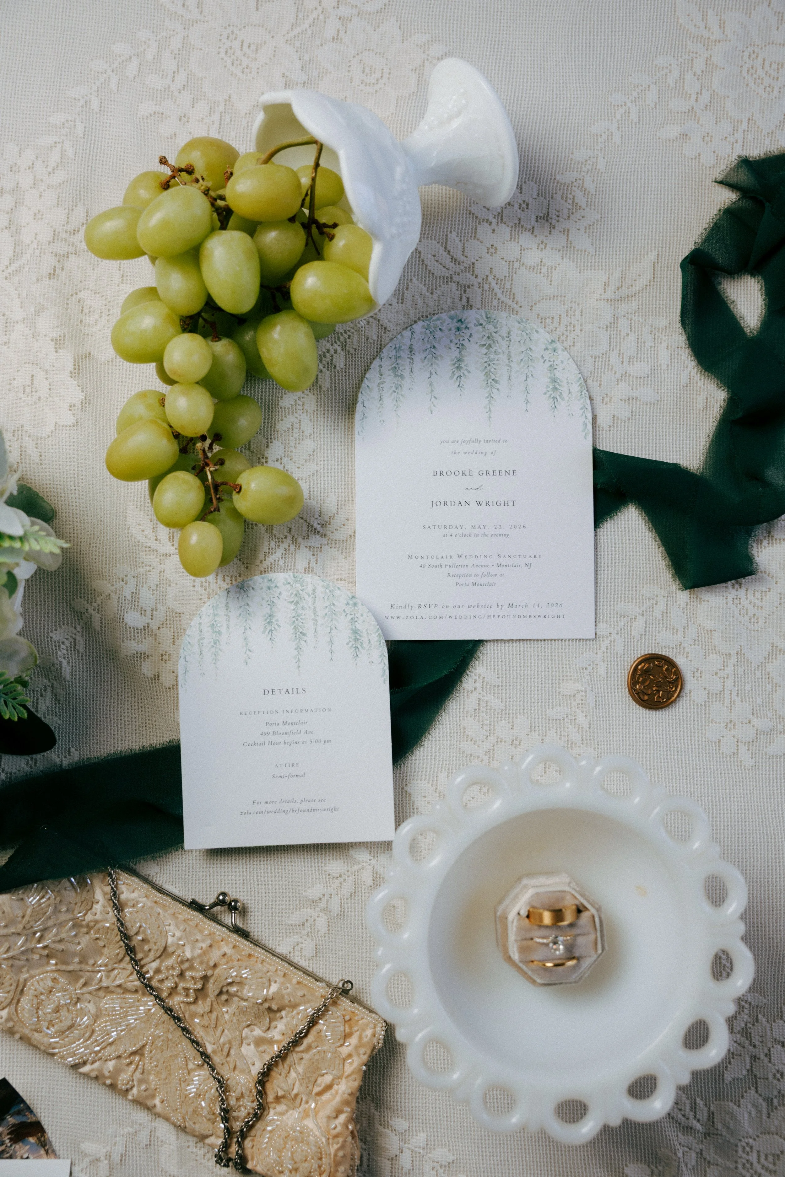 Wedding invitation cards with greenery design, green grapes on a vine, a white ceramic goblet, a gold ring on a small hexagonal ring holder, a lace clutch with a black chain, and a white ceramic plate with small holes around the edge, all arranged on a lace tablecloth.