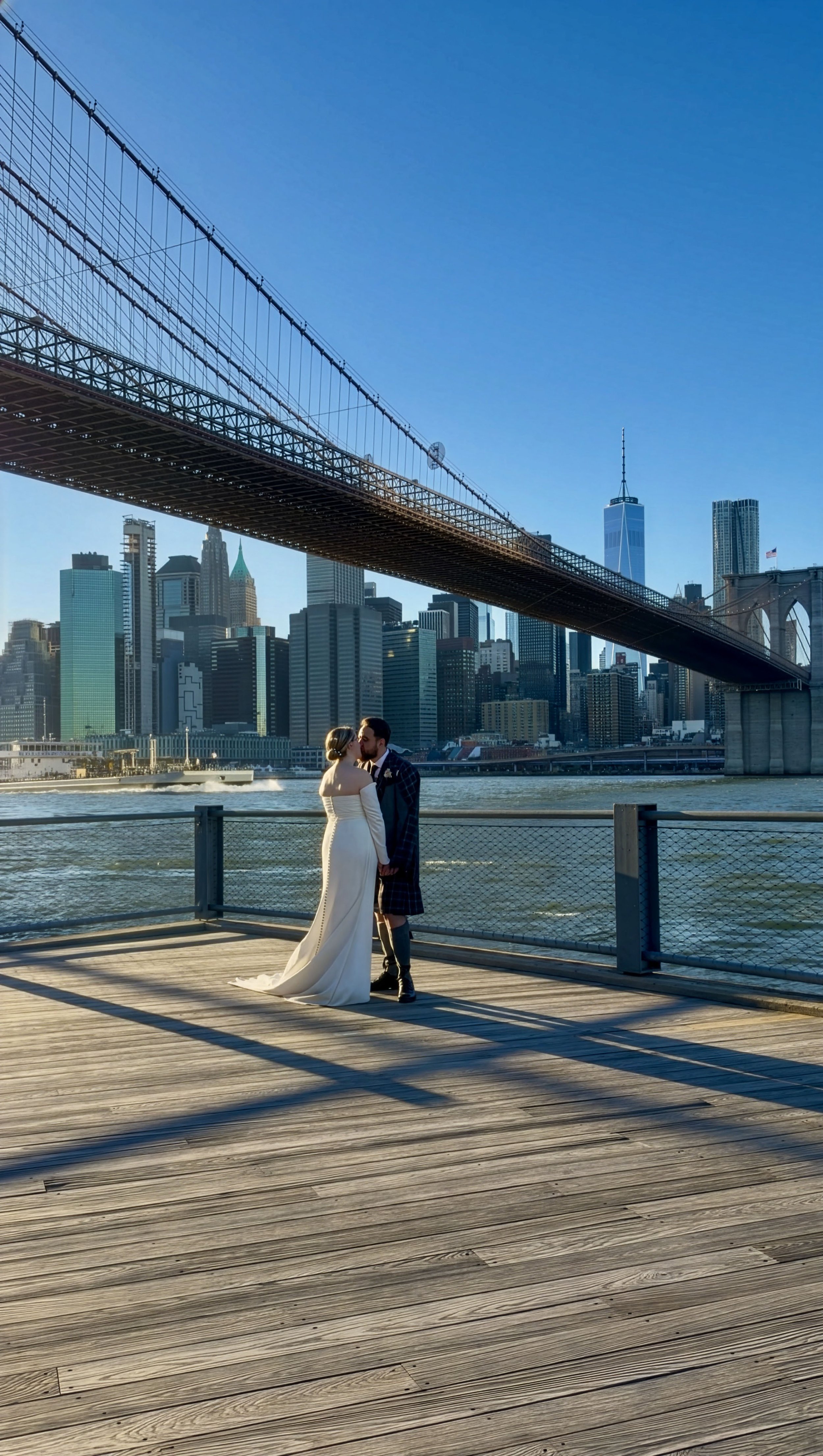 Couple dressed in wedding attire sharing a kiss on a wooden pier over the water, with the New York City skyline and Brooklyn Bridge in the background.