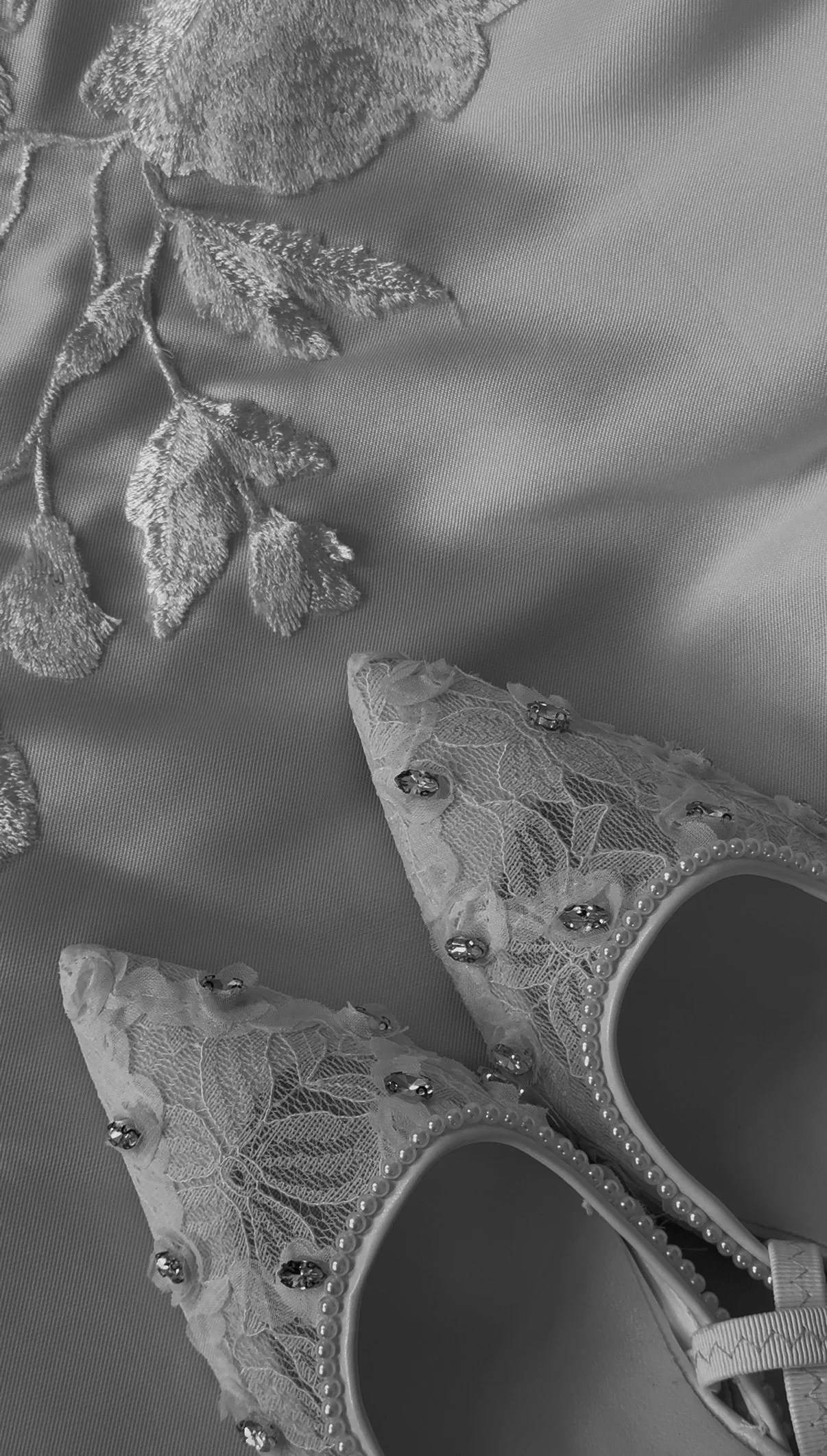 A pair of lace slippers decorated with small beads and pearls, placed on silky fabric with embroidered leaves.