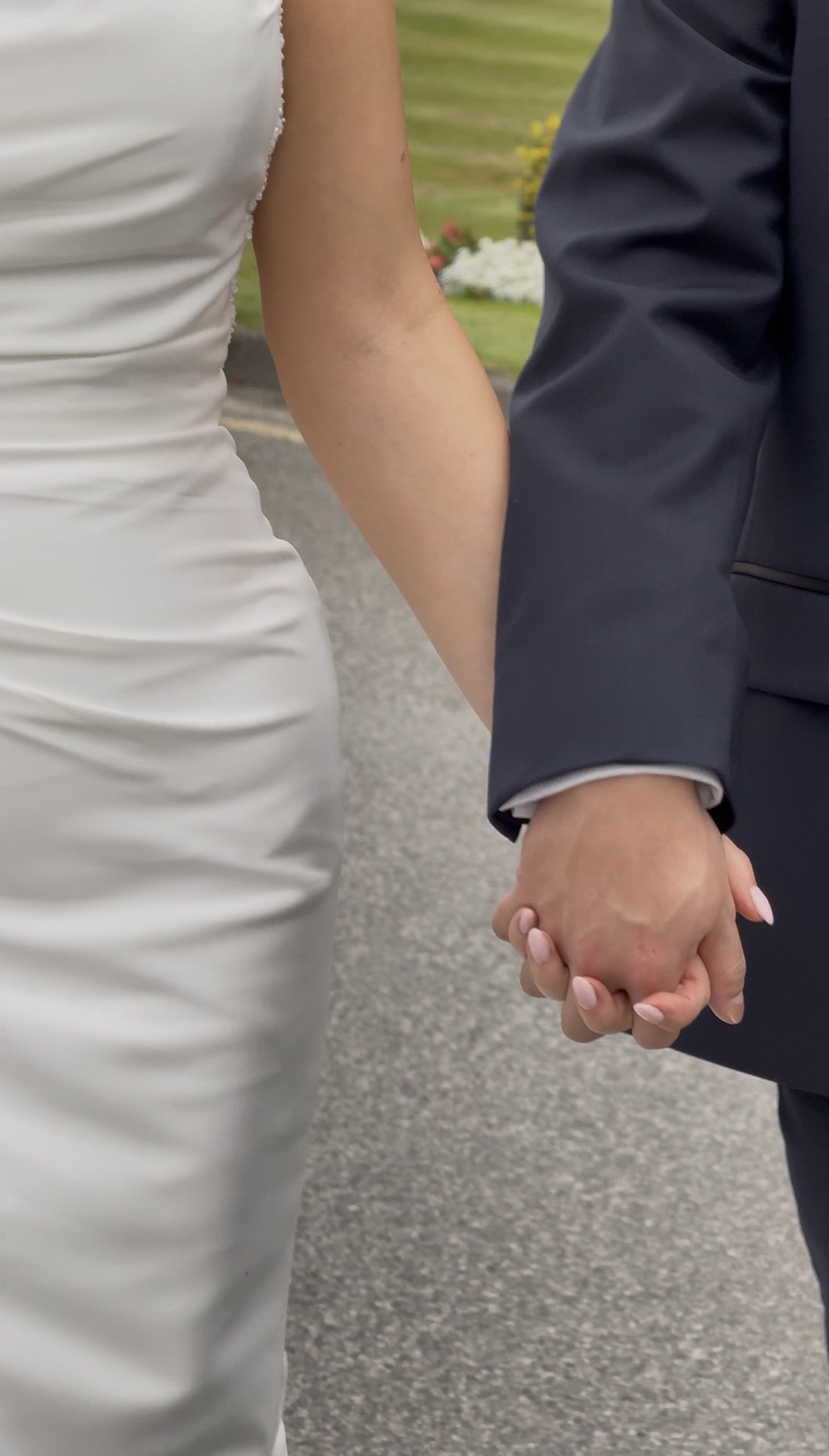 Close-up of a couple holding hands during a wedding ceremony, with a bride in a white dress and a groom in a dark suit, standing outdoors with a garden and flowers in the background.