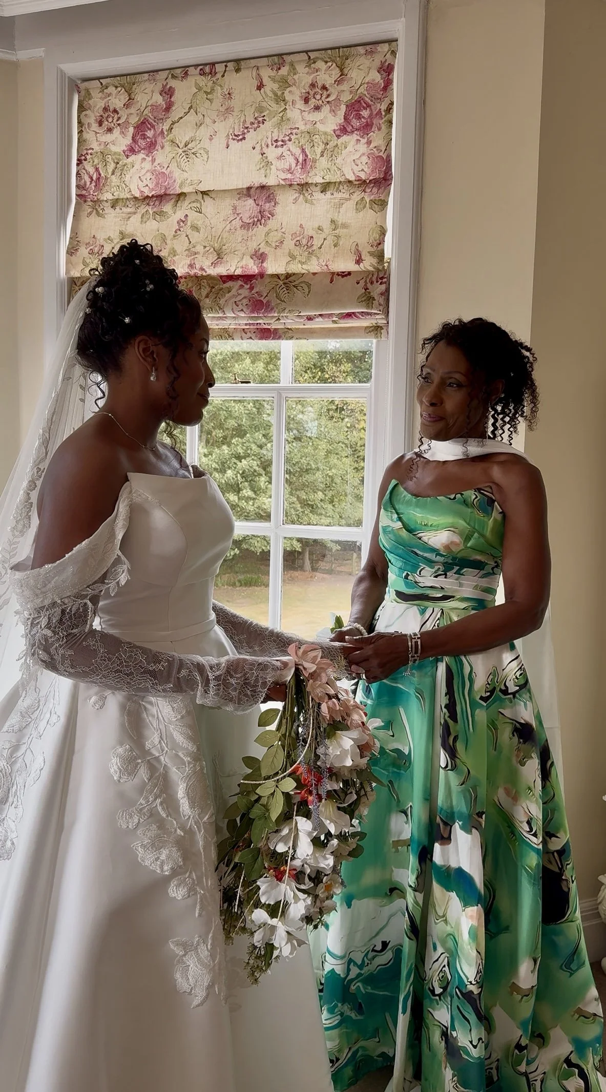 A bride in a white wedding dress holding a bouquet standing next to a woman in a green and white patterned dress, both facing each other by a window with floral curtains.