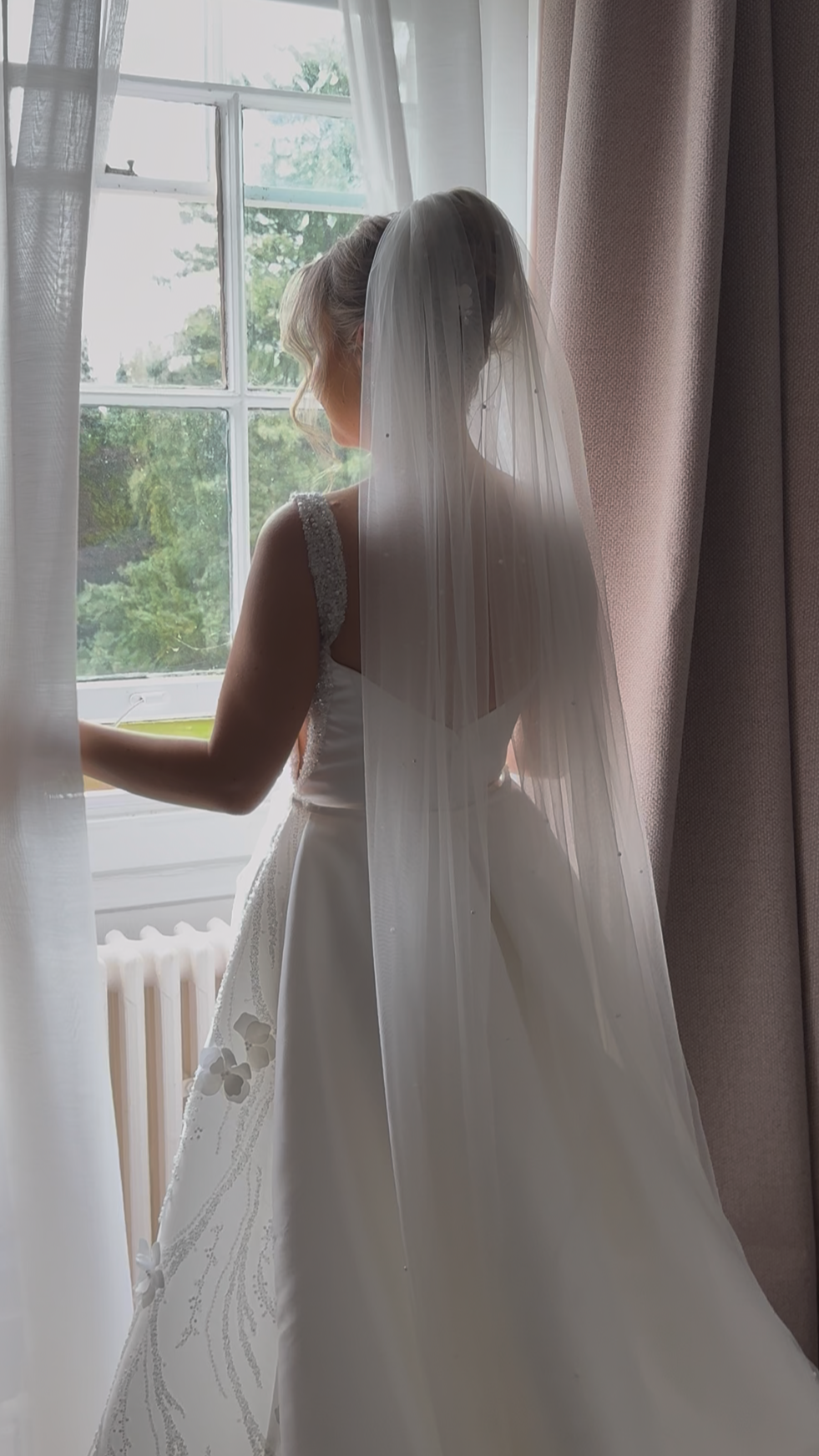 A bride in a white wedding gown with a long veil looks out the window on her wedding day.