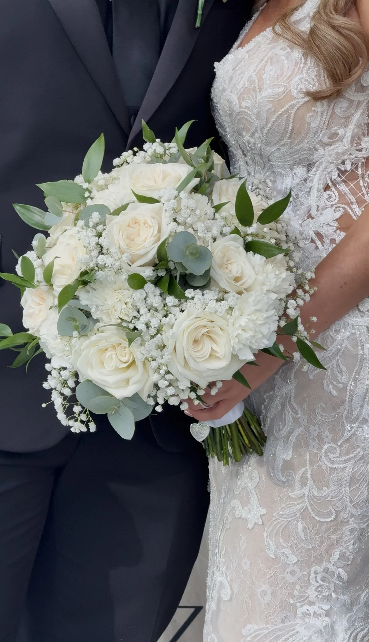 Close-up of a bride and groom holding a white wedding bouquet, with the bride's lace wedding dress visible.