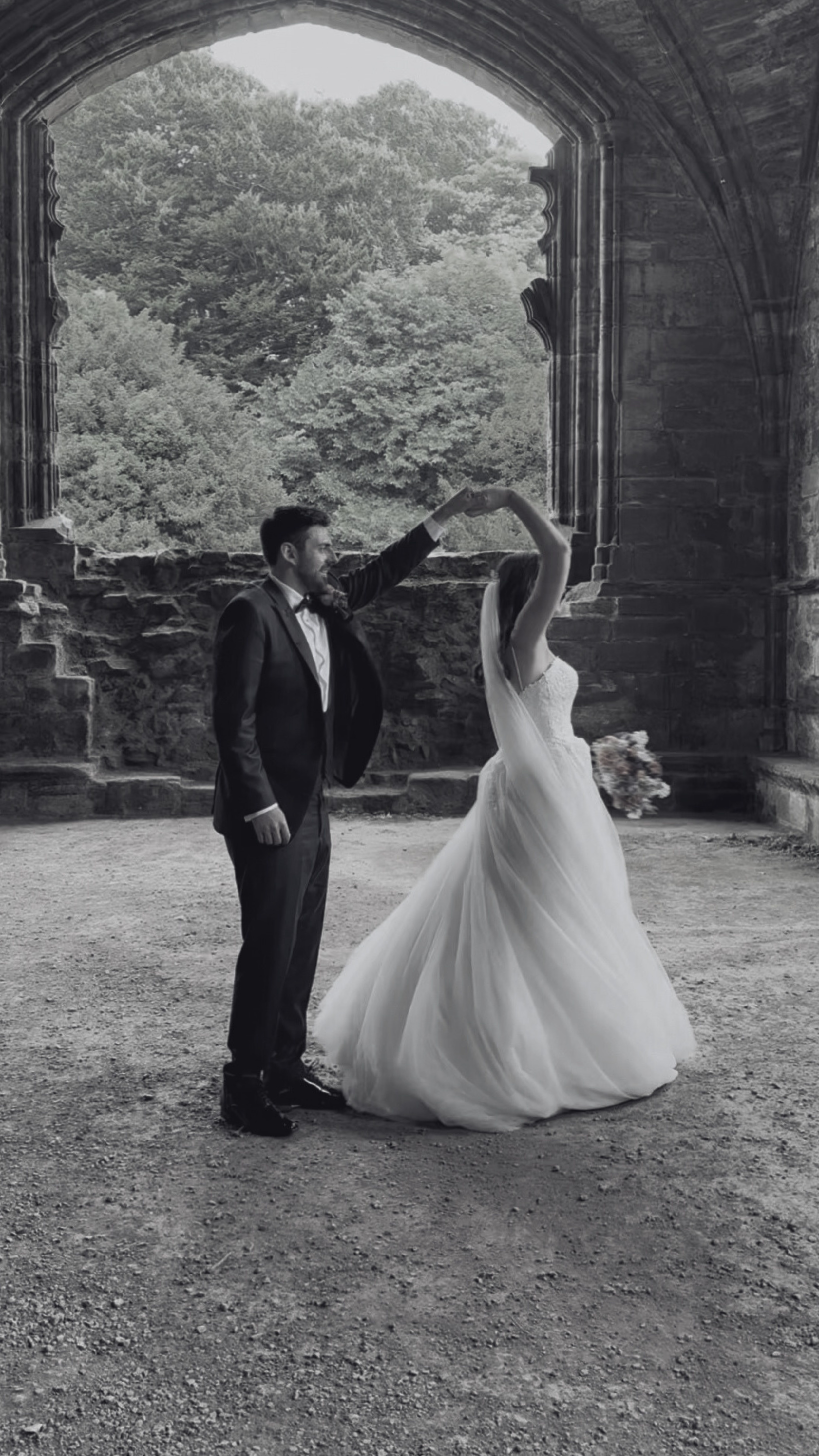 A black and white photo of a bride and groom dancing inside a stone building with large arched window, overlooking a scenic view of trees. The groom is wearing a black suit and the bride is in a flowing white wedding gown, holding a bouquet in her le