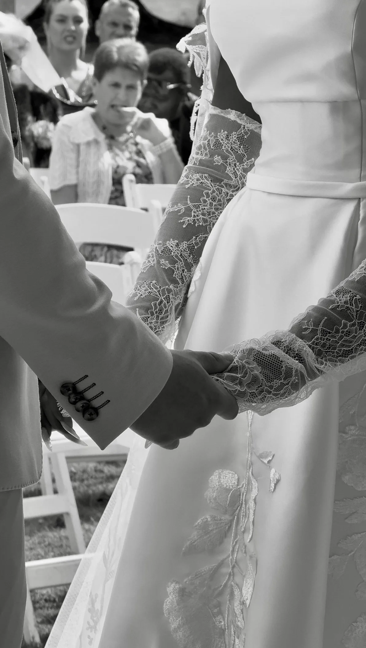A close-up black and white photo of a wedding ceremony showing the bride and groom holding hands. The bride wears a satin dress with lace sleeves and lace detail along the skirt. The groom wears a suit with buttons on the sleeve. In the background, g