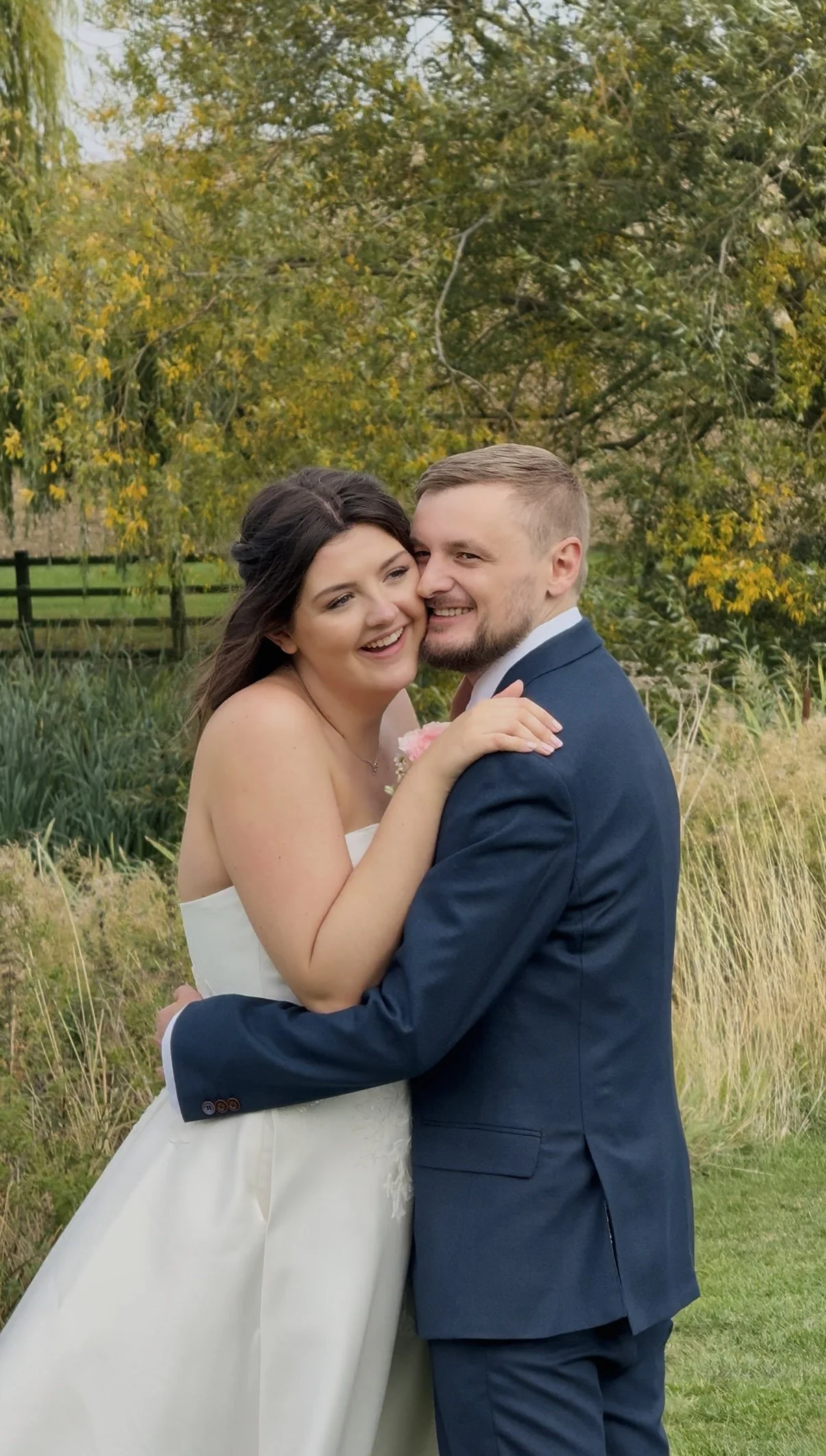 A smiling bride and groom embracing outdoors on their wedding day, with trees and grass in the background.