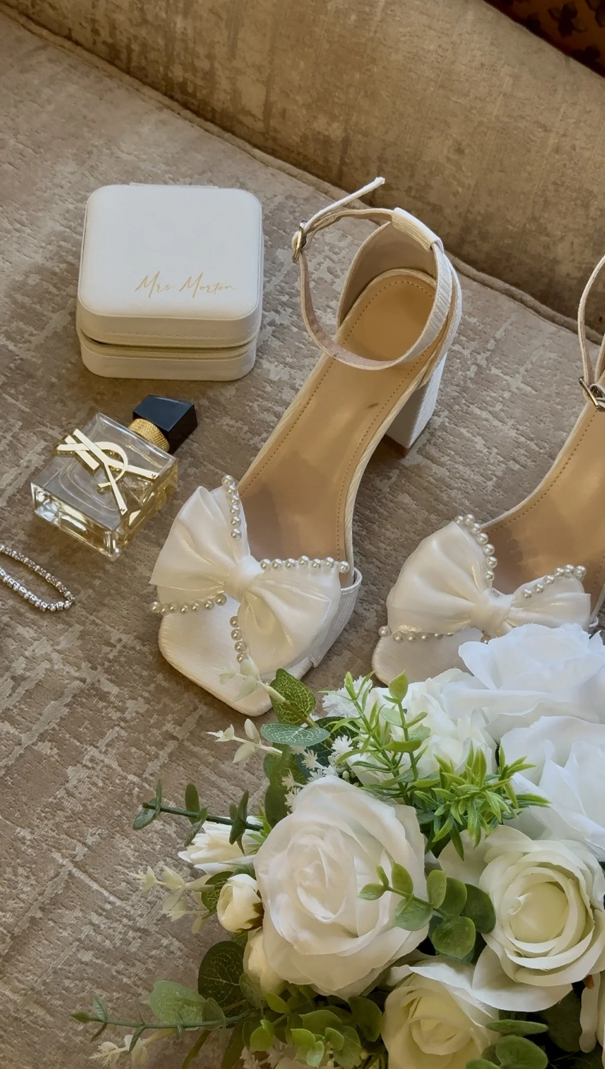 Bridal accessories including a pair of white high-heeled shoes with pearl bows, a bouquet of white roses and greenery, a small white jewelry box labeled 'Mrs. Morton,' a bottle of perfume, a jewelry piece, and a white clutch bag placed on a beige tex