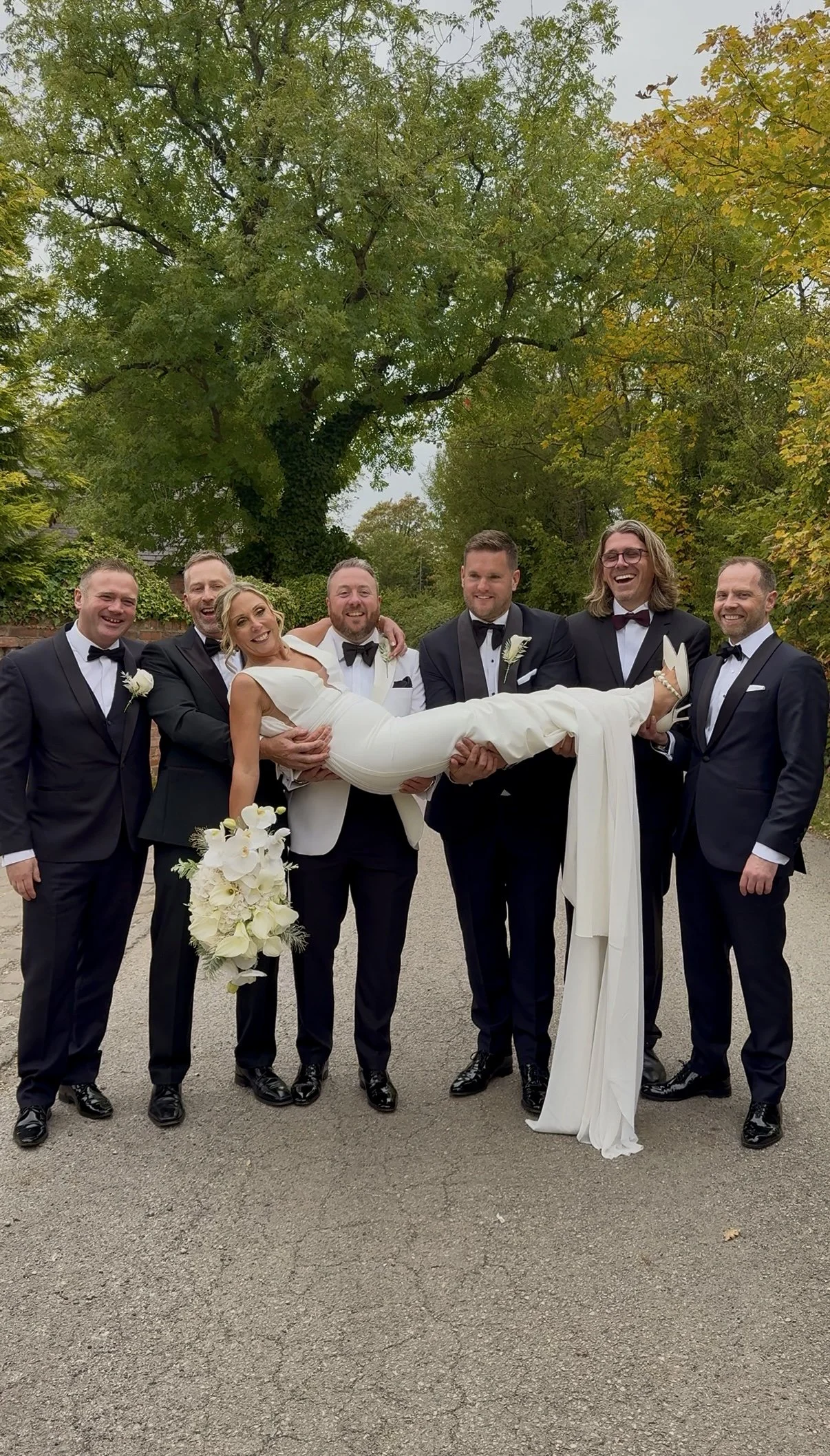 Group of seven men in tuxedos lifting a bride in a white wedding dress outdoors during the daytime, smiles, trees with green and yellow leaves in the background.