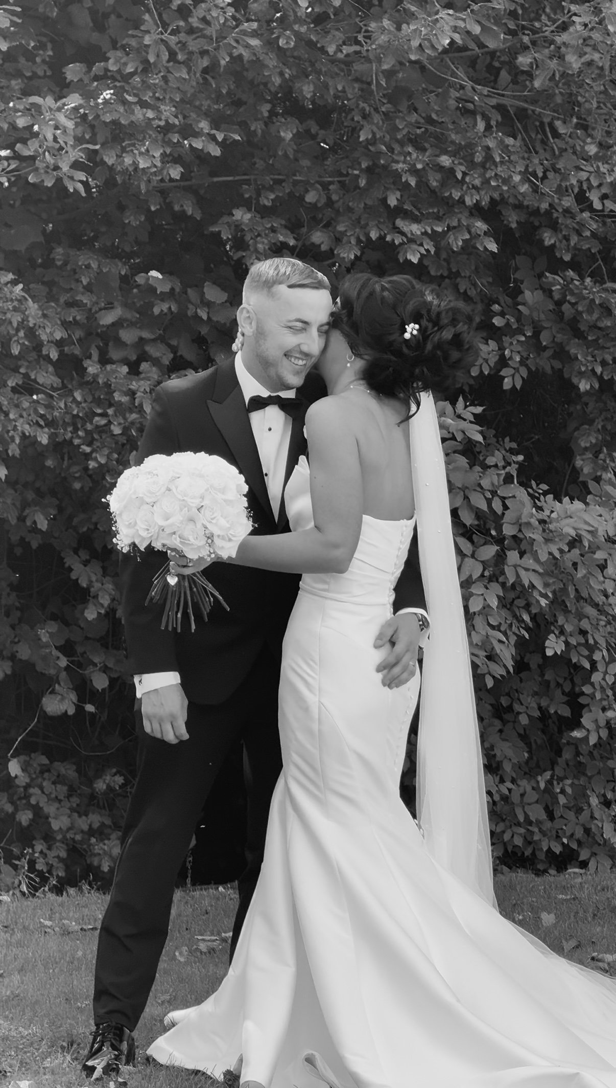 A black-and-white photograph of a newly married couple in wedding attire. The groom, in a tuxedo, and the bride, in a strapless wedding gown with a long train and veil, are happily embracing outdoors in front of lush foliage.