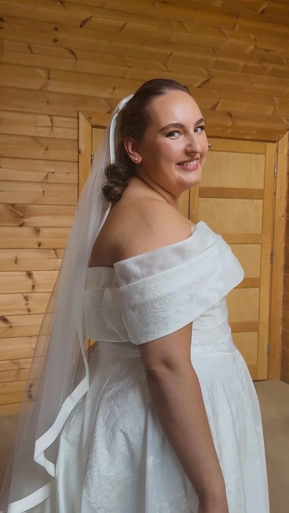 A smiling bride in a white wedding dress with off-the-shoulder sleeves, standing in a wooden room, wearing a veil.